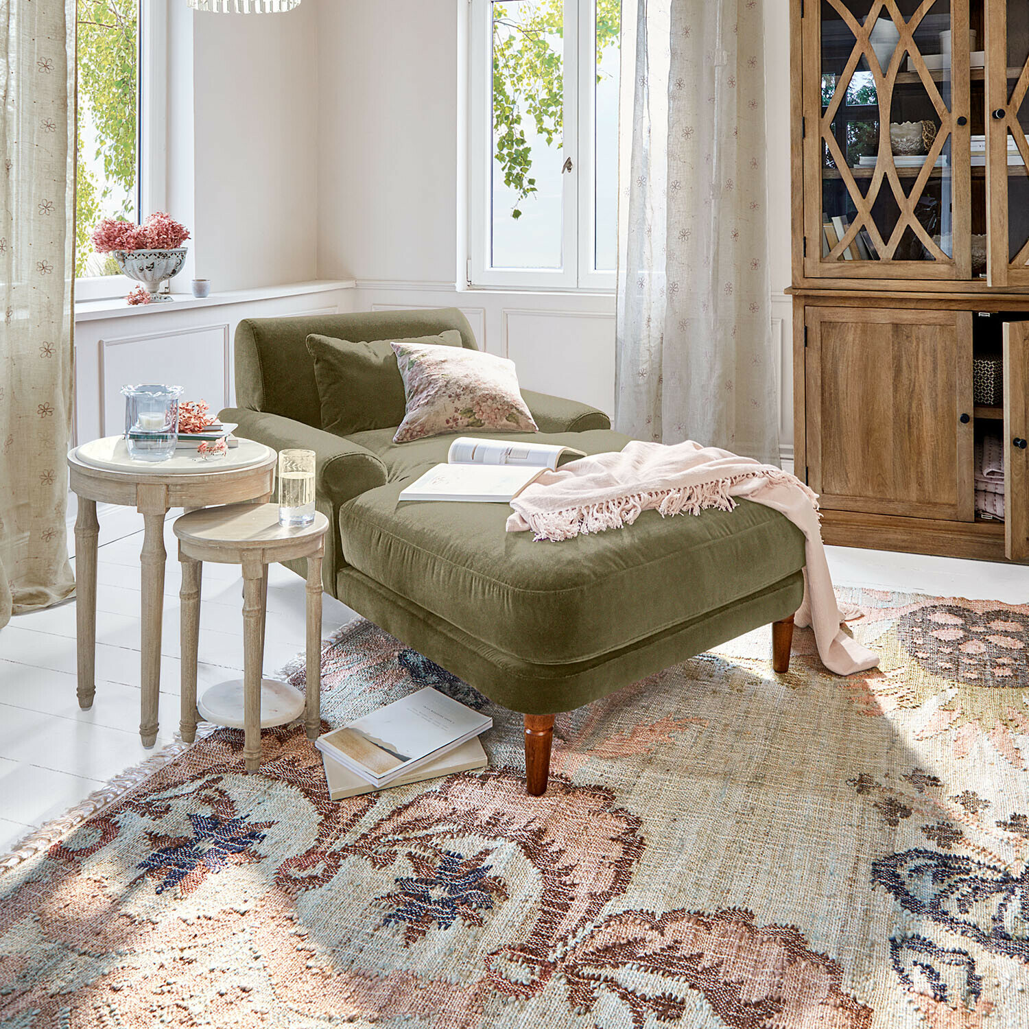 Groene fluwelen chaise longue met kussens en roze deken in lichte woonkamer met houten meubels