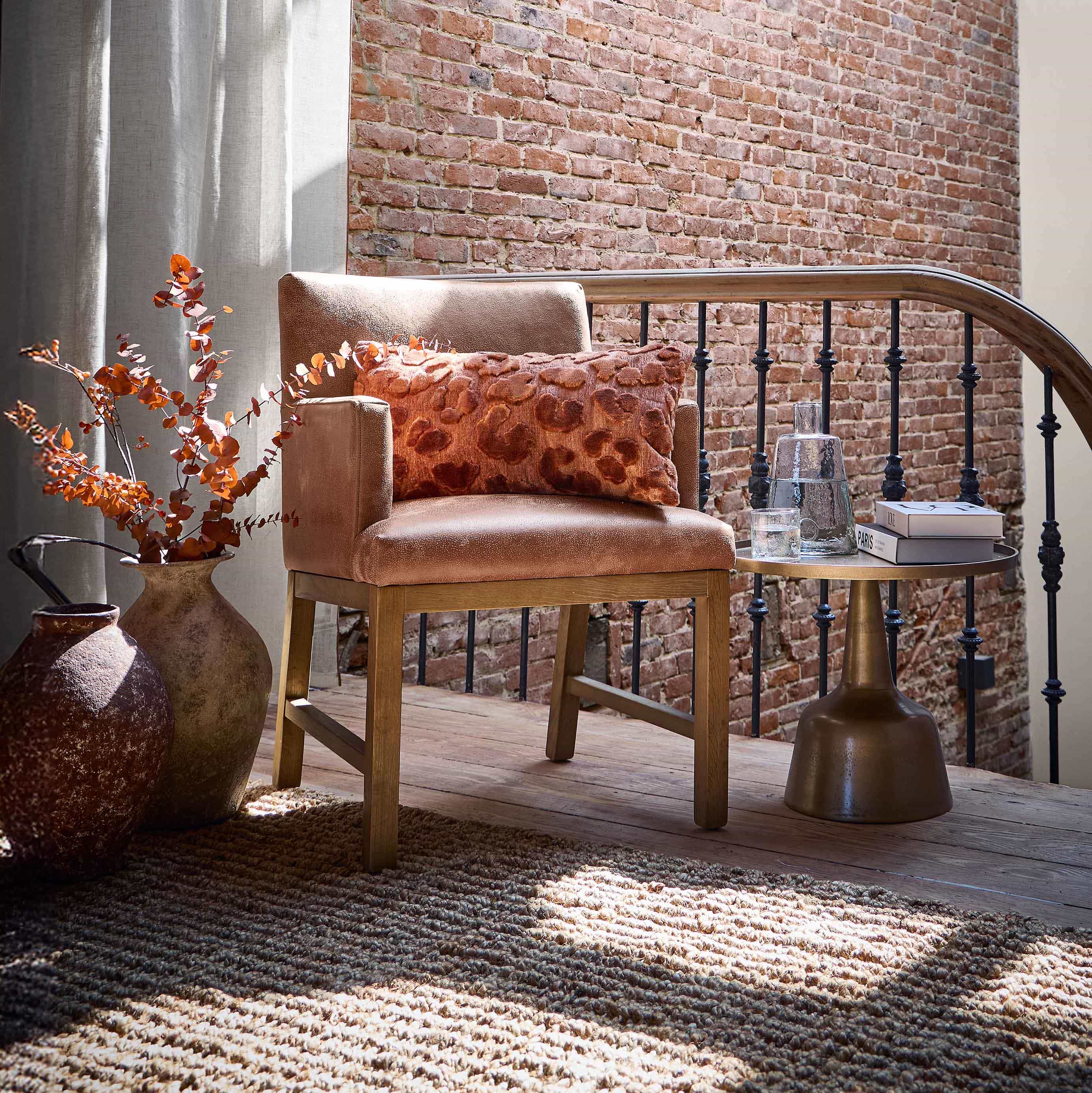 Beige fauteuil met houten onderstel en oranje kussen voor een bakstenen muur en gordijnen