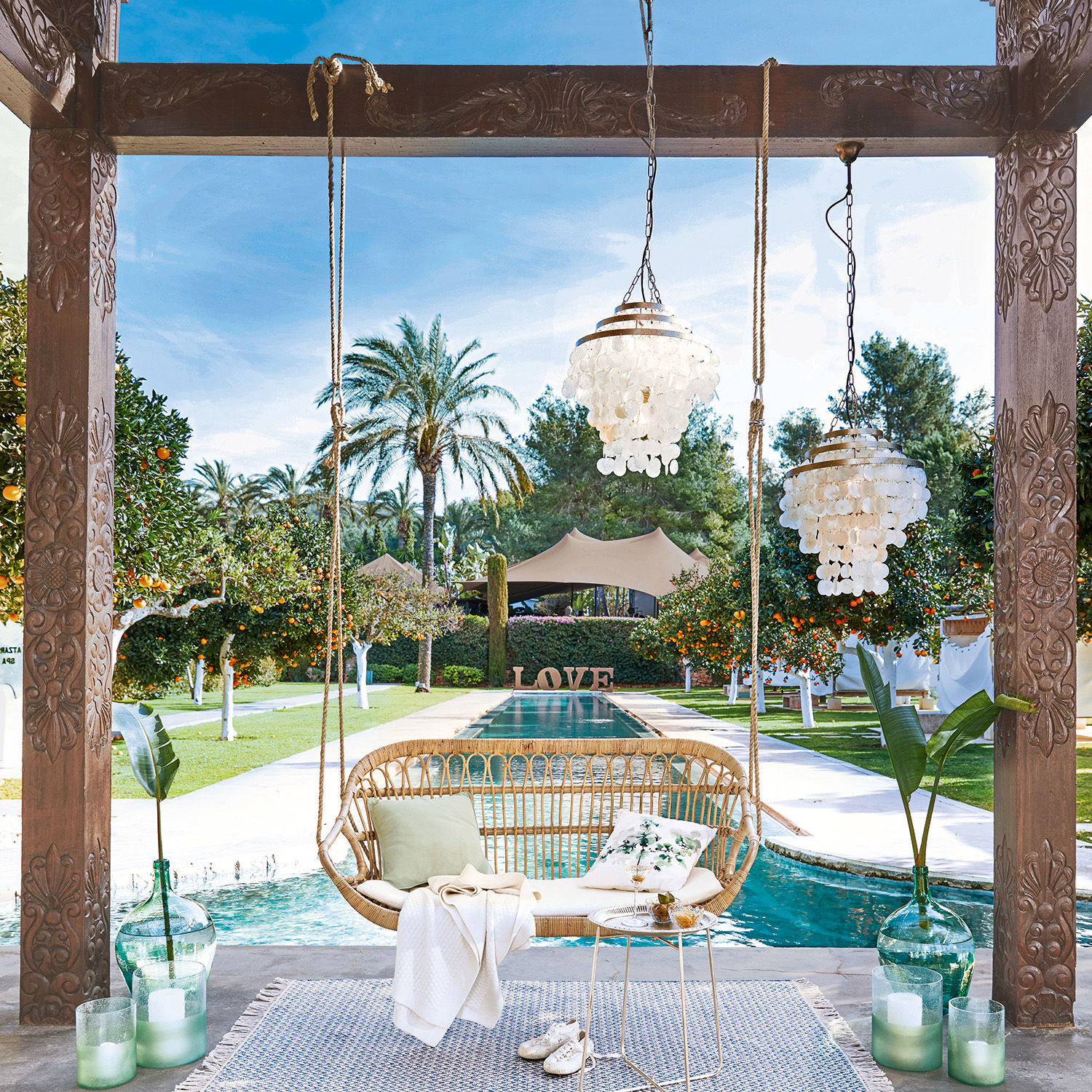 Rotan schommel met kussens en deken bij zwembad in tuin met palmbomen en sinaasappelbomen