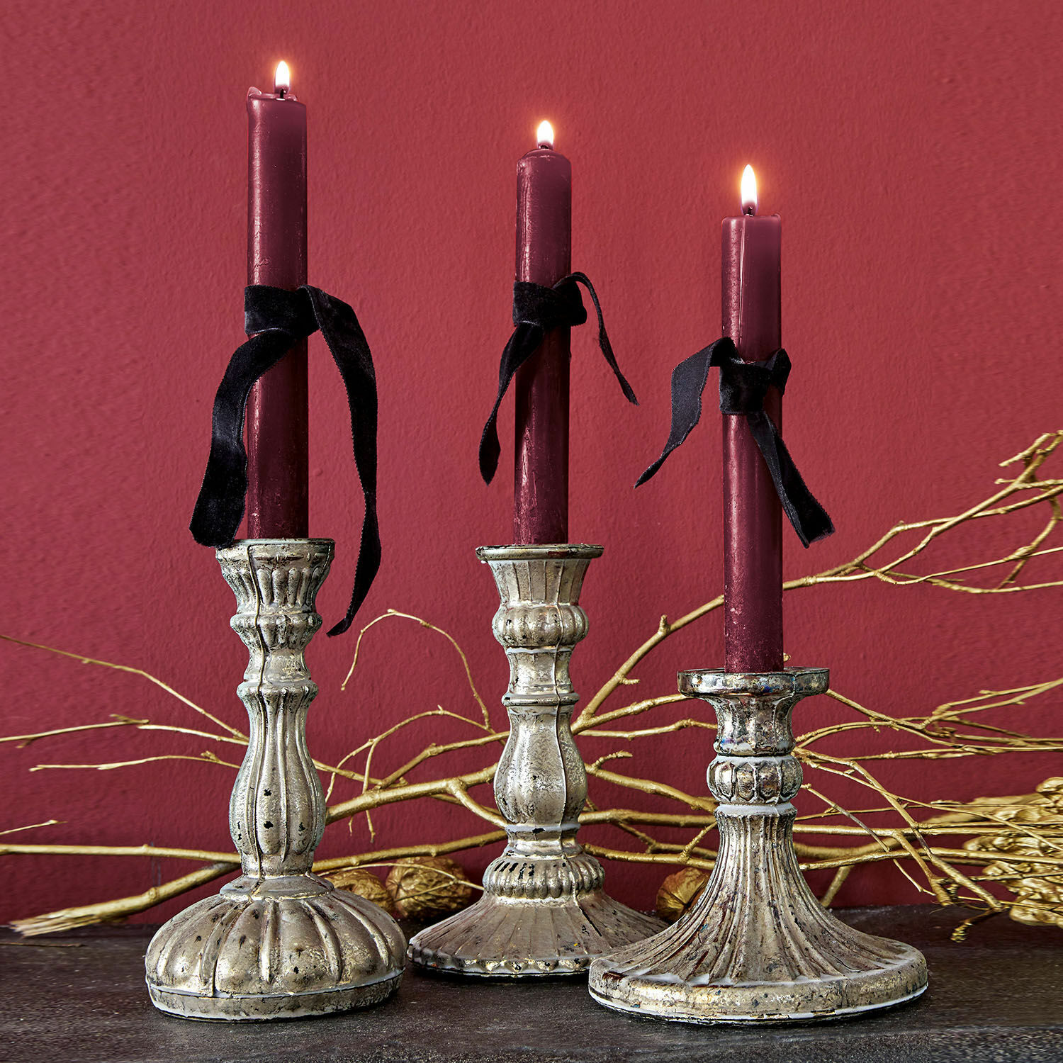 Drie decoratieve metalen kandelaars met bordeauxrode kaarsen en zwarte linten op een donkere achtergrond