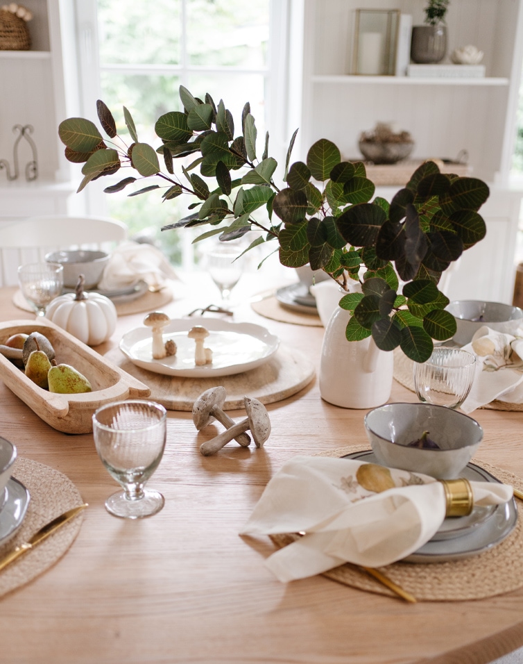 Herfsttafel van licht hout met groene bladeren, decoratieve paddenstoelen en stijlvolle servies voor diner