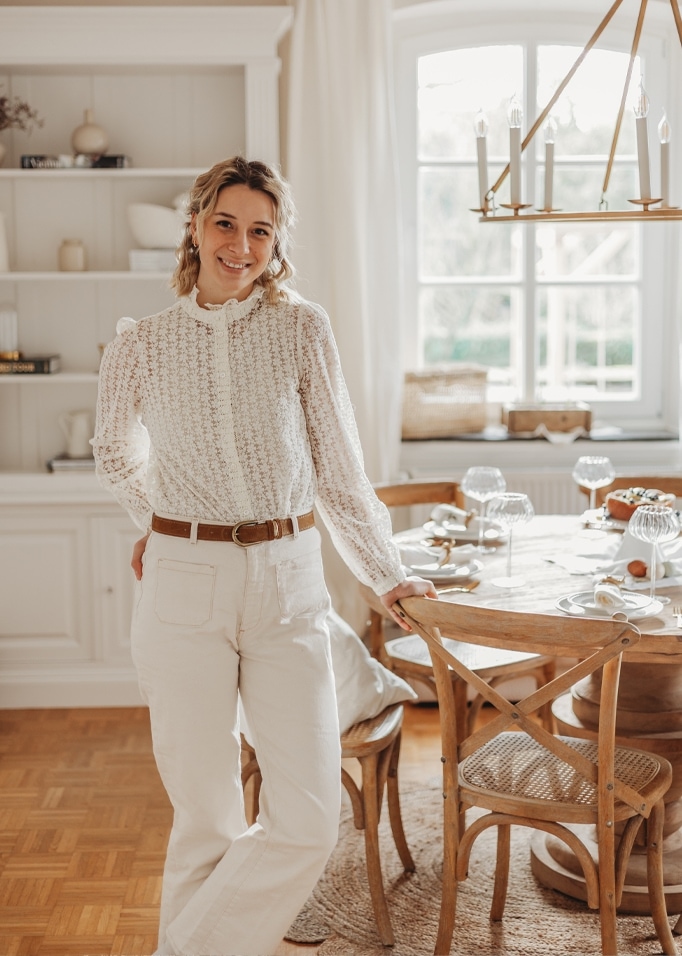 Vrouw in witte kanten blouse en lichte broek bij houten tafel in lichte kamer.