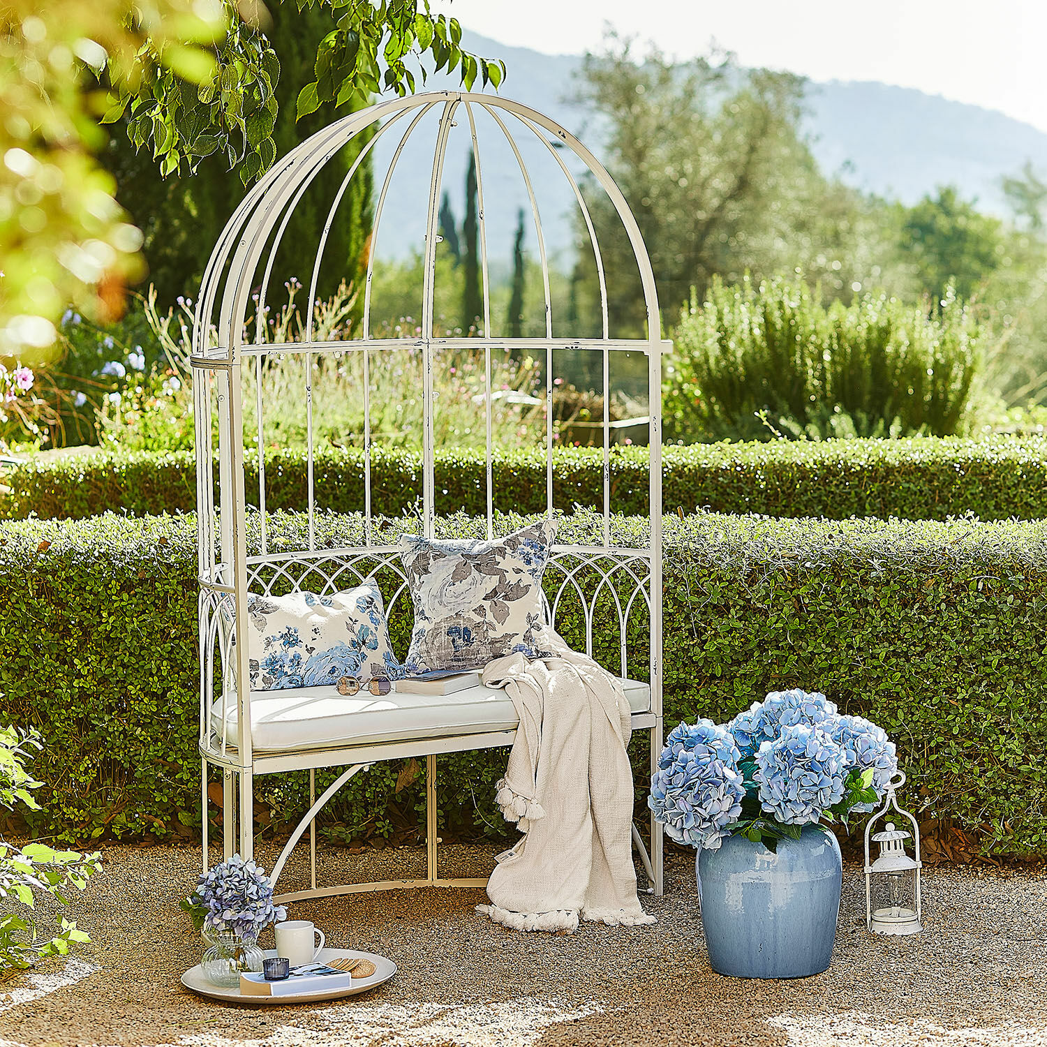 Witte metalen tuinbank met kussens en deken, versierd met blauwe hortensia bloemen