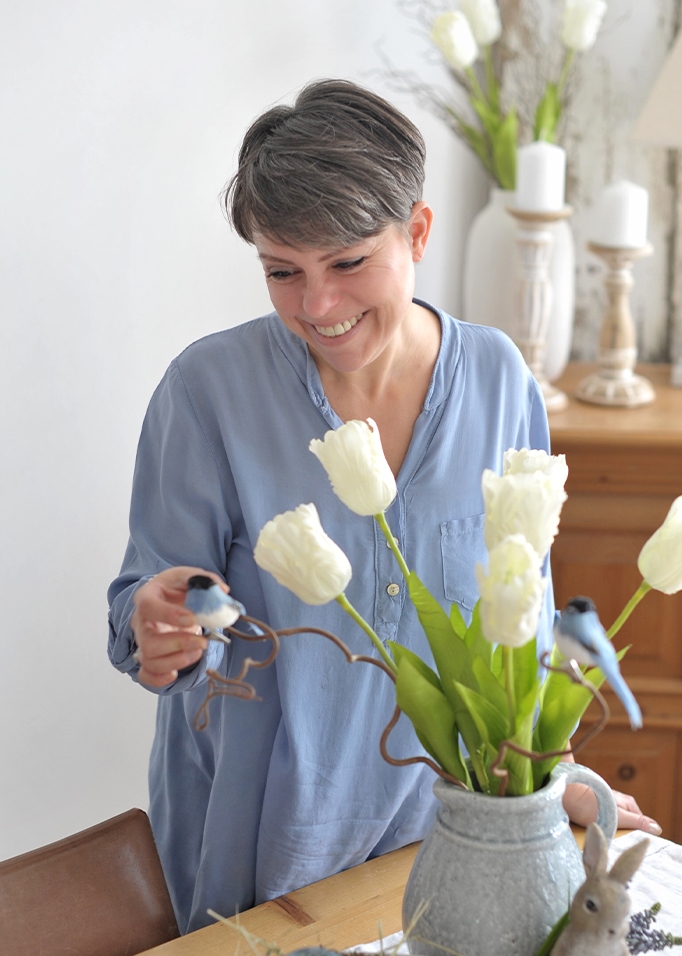 Vrouw in blauw overhemd decoreert tafel met grijze keramische vaas, witte tulpen en decoratieve vogels