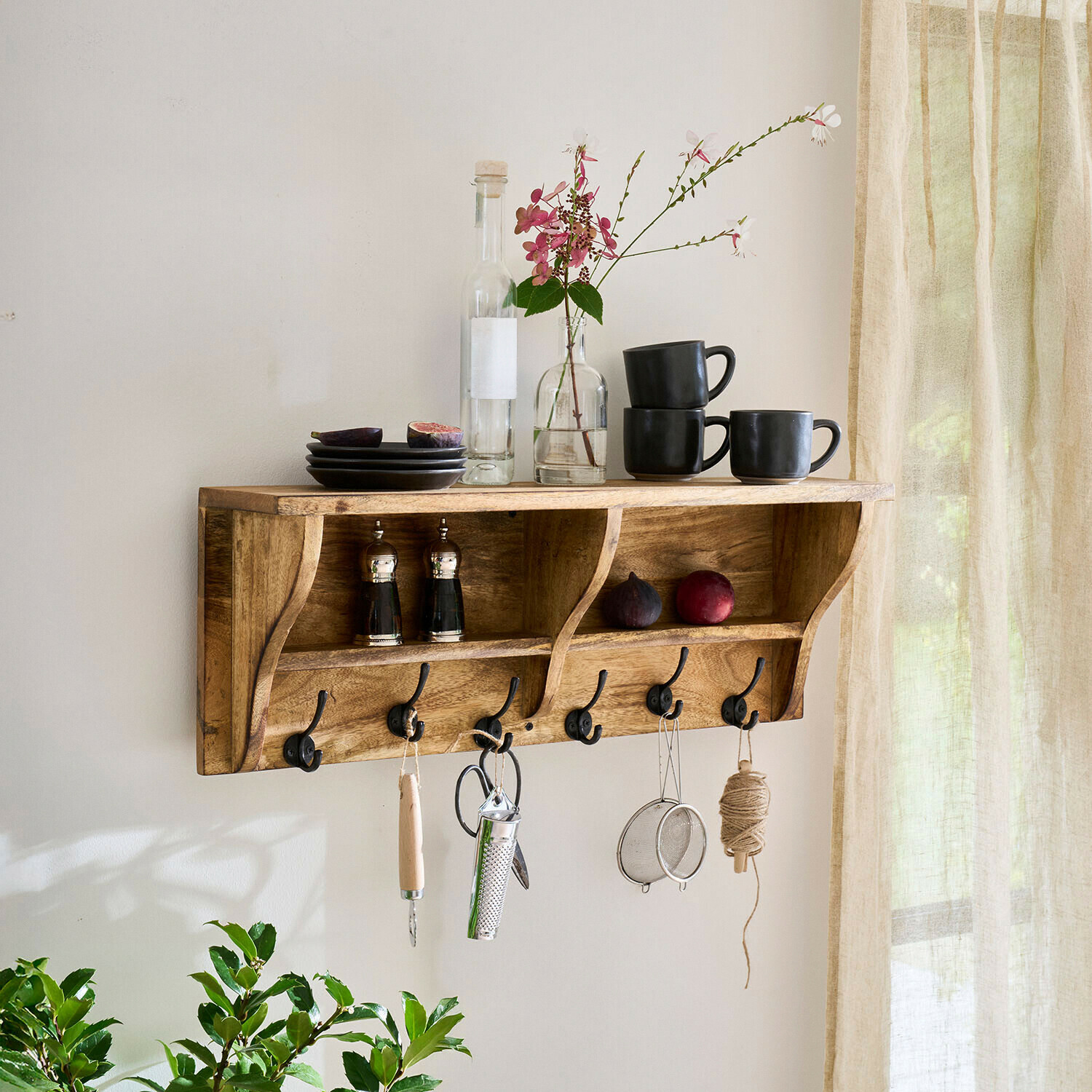 Houten wandplank met haken, zwarte mokken, borden en bloemenarrangement in lichte kamer