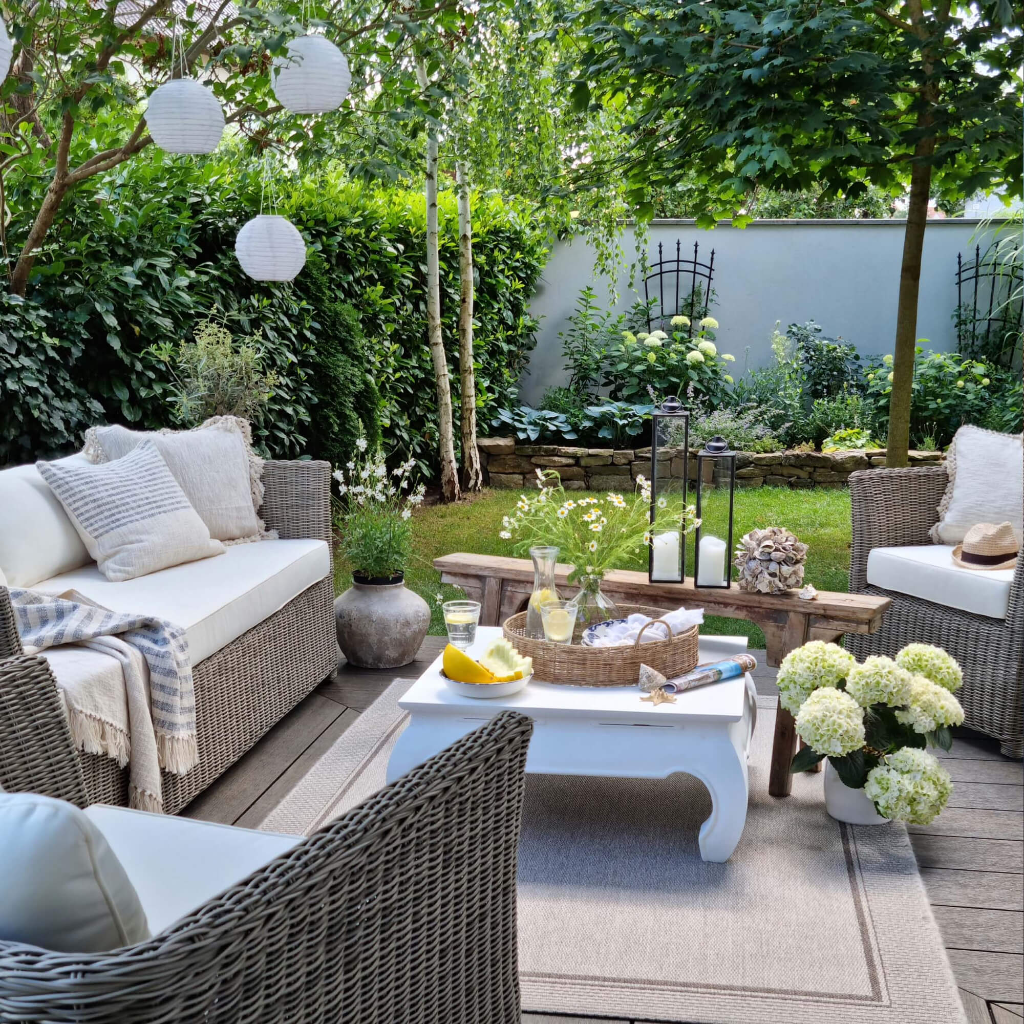 Gezellige tuinhoek met grijze rotanmeubels en witte kussens, decoraties en groene planten.