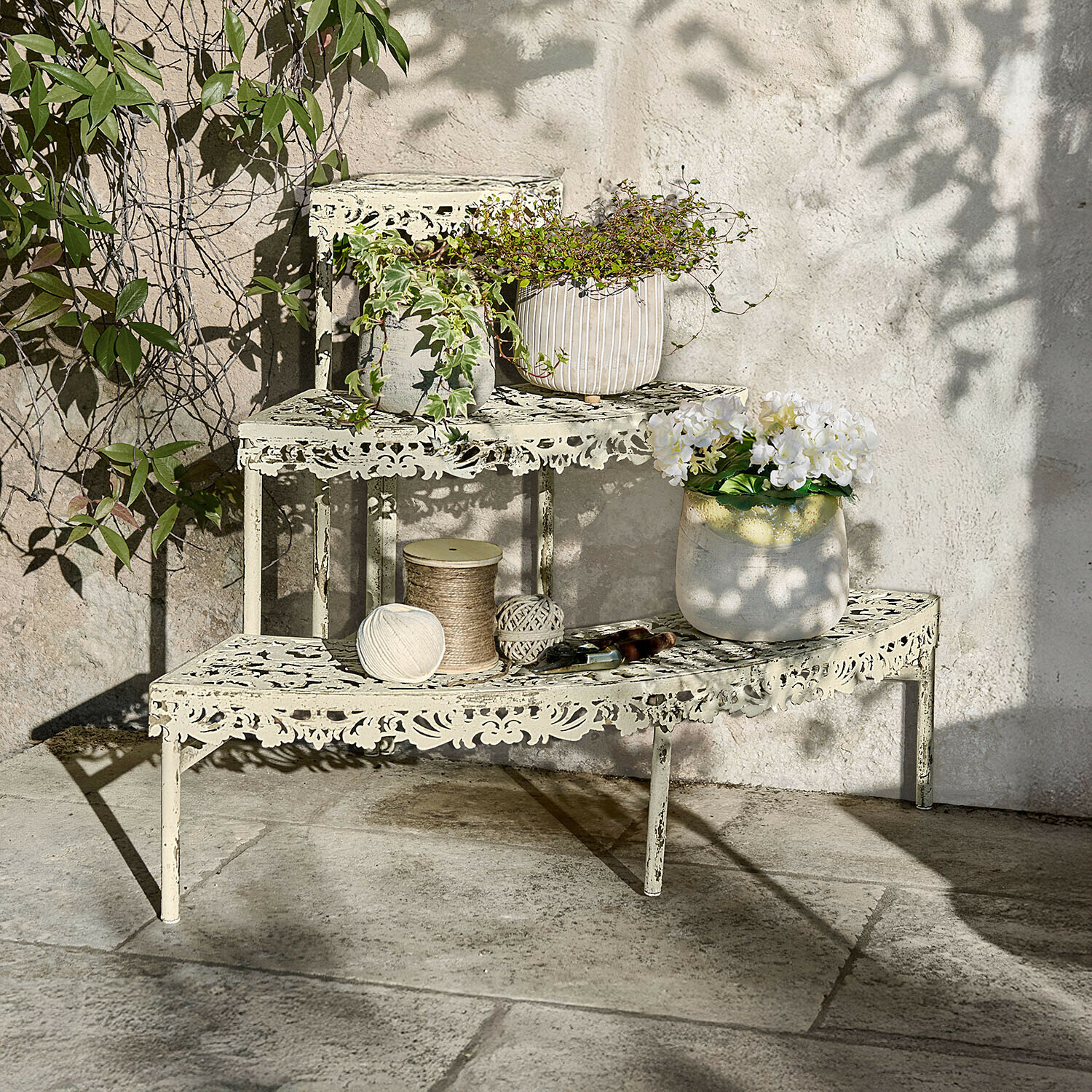 Crèmekleurige metalen etagère met opengewerkt patroon, planten en tuingereedschap op stenen terras