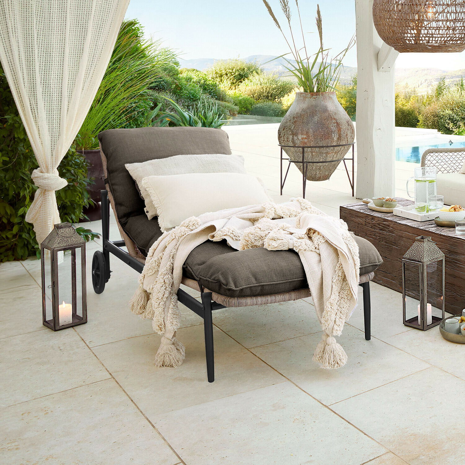 Tuinligstoel met donkergrijze stof en crèmekleurige deken met franjes op terras met tuinzicht