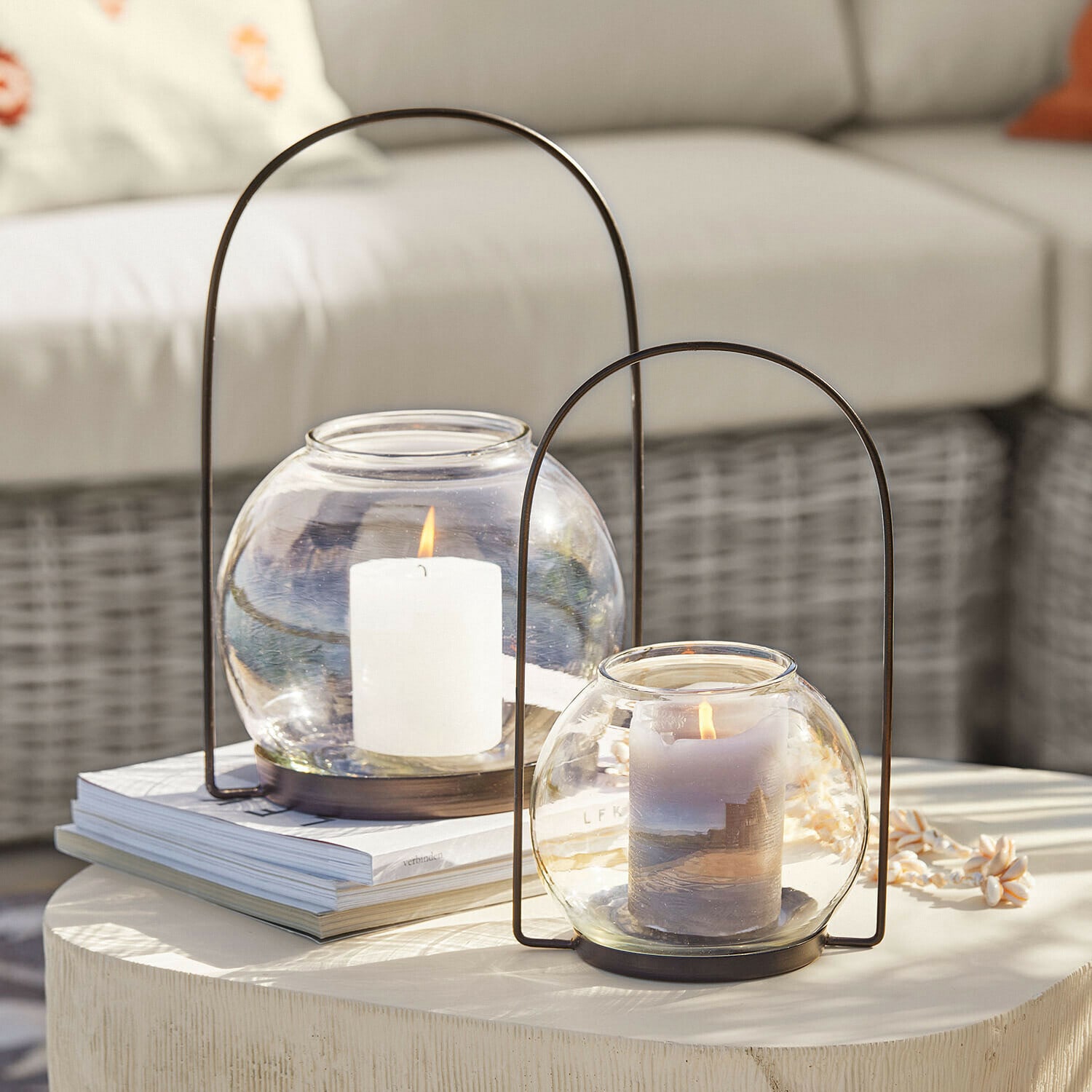 Twee glazen lantaarns met metalen handvatten en brandende kaarsen op een lichte decoratieve tafel