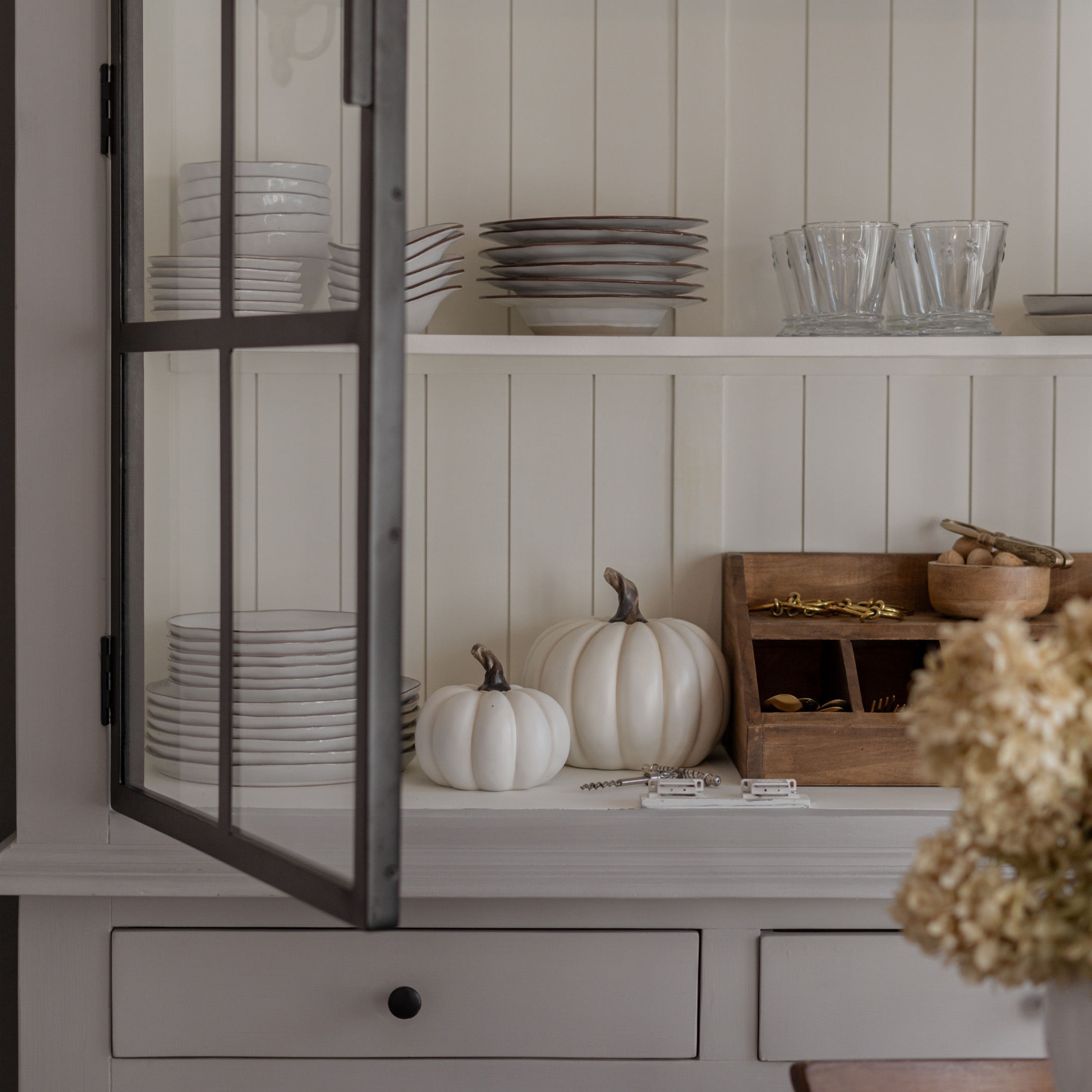 Witte keramische borden en glazen op plank met decoratieve witte pompoenen en houten organizer
