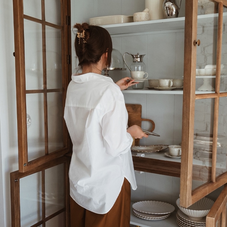 Vrouw in wit overhemd pakt porseleinen kopje uit houten vitrinekast met servies