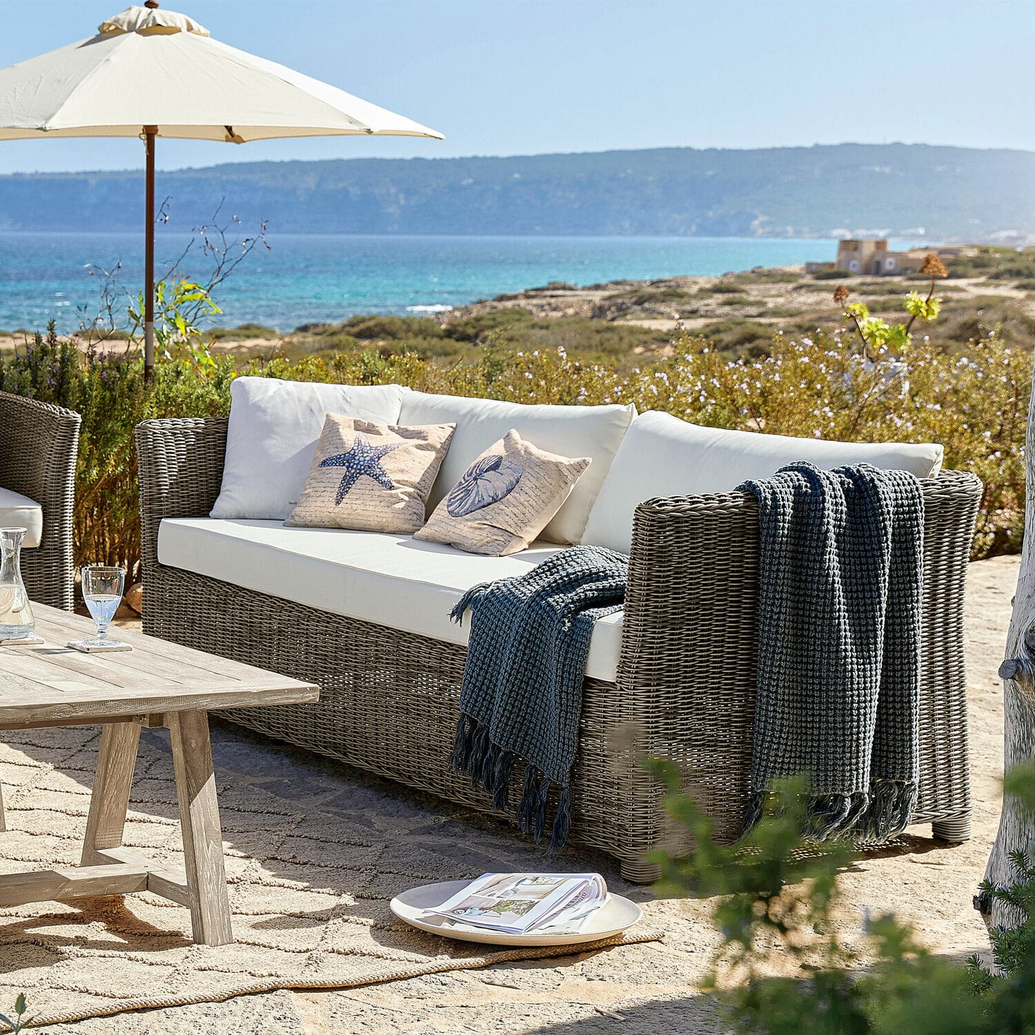 Tuinbank van grijs rattan met witte kussens en decoratieve hoezen met zee-thema voor terras