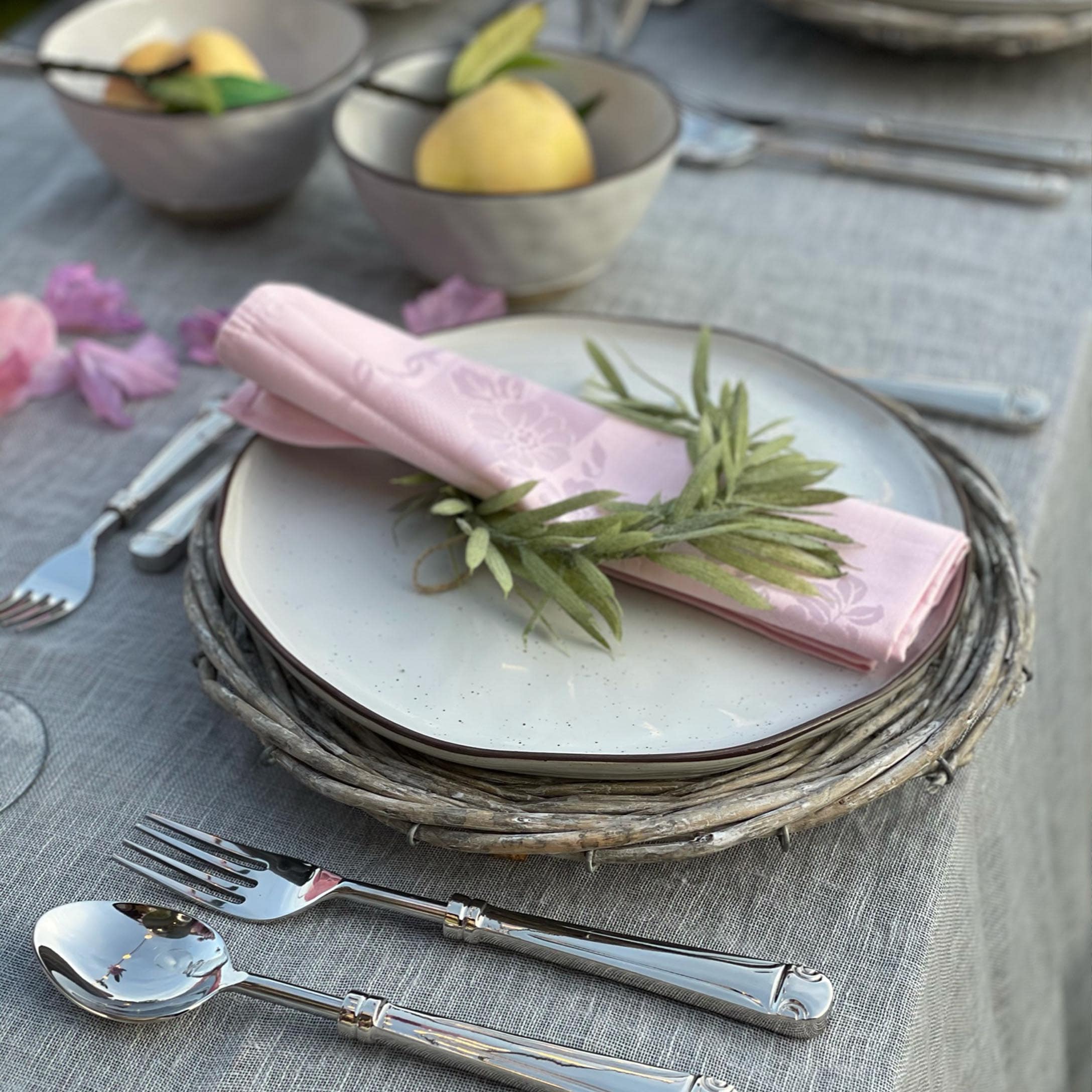 Elegante tafelsetting met wit bord op rieten onderzetter en roze servet met groene bladeren