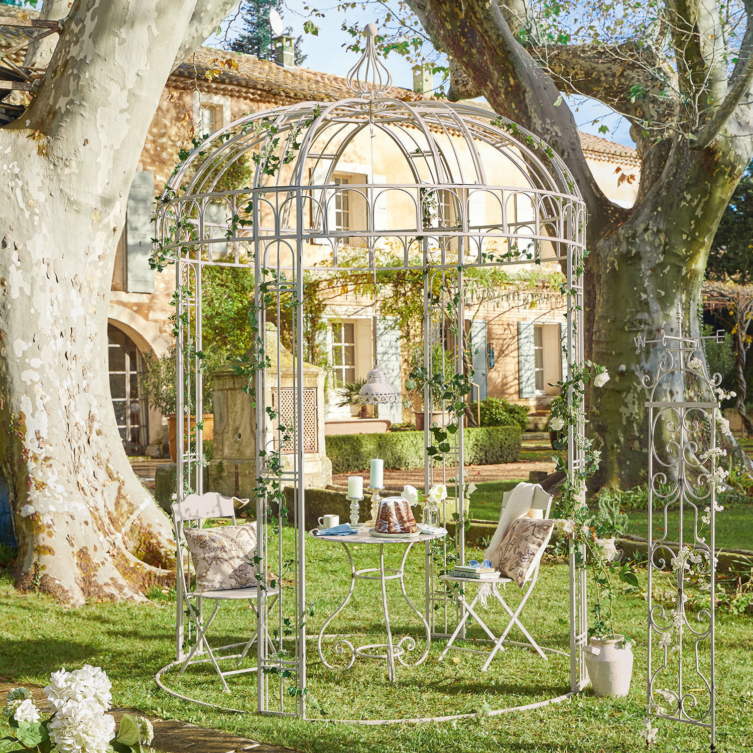 Paviljoen Iolanda Witte metalen tuinprieel met stoelen en tafel op gras, versierd met groene klimop