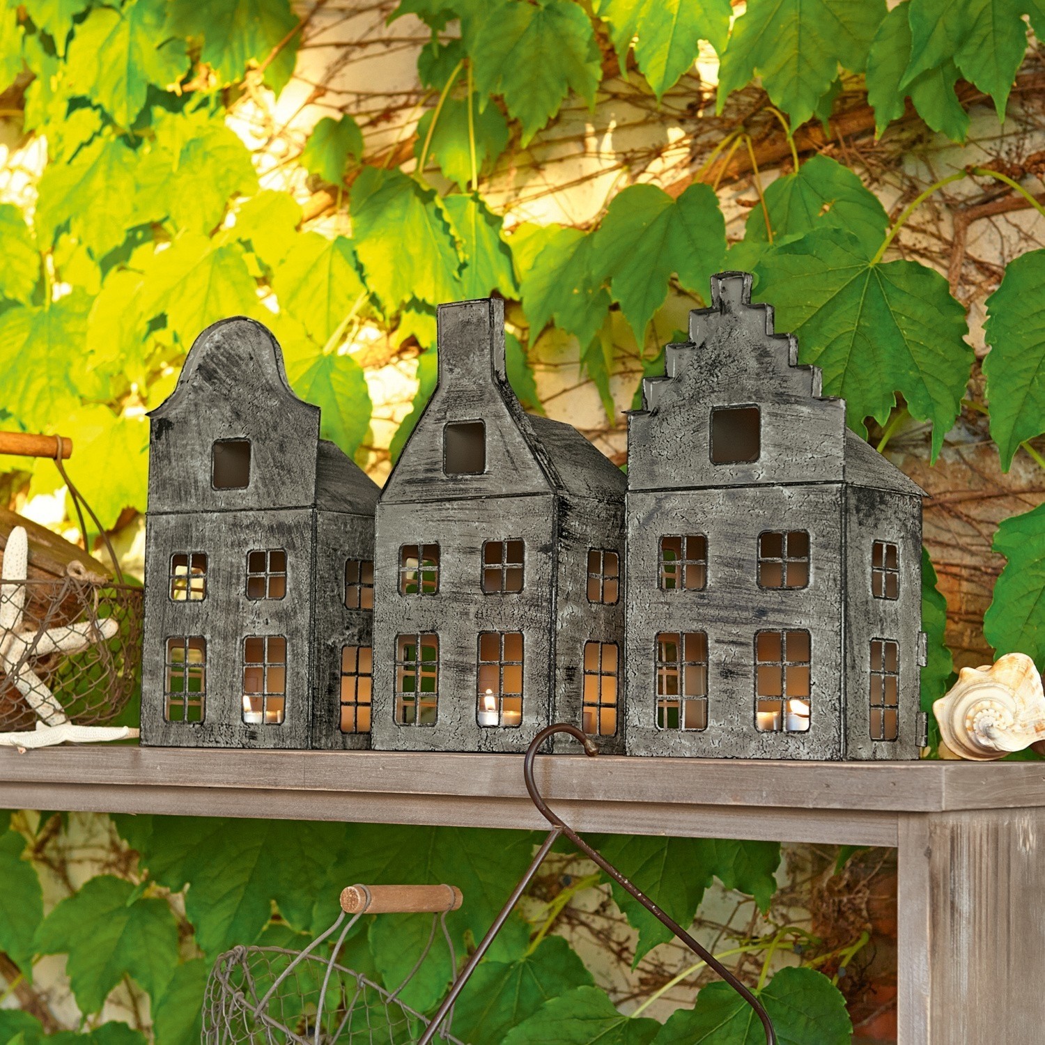 Grijze metalen lantaarns in huisvorm op houten plank met groene bladeren op de achtergrond