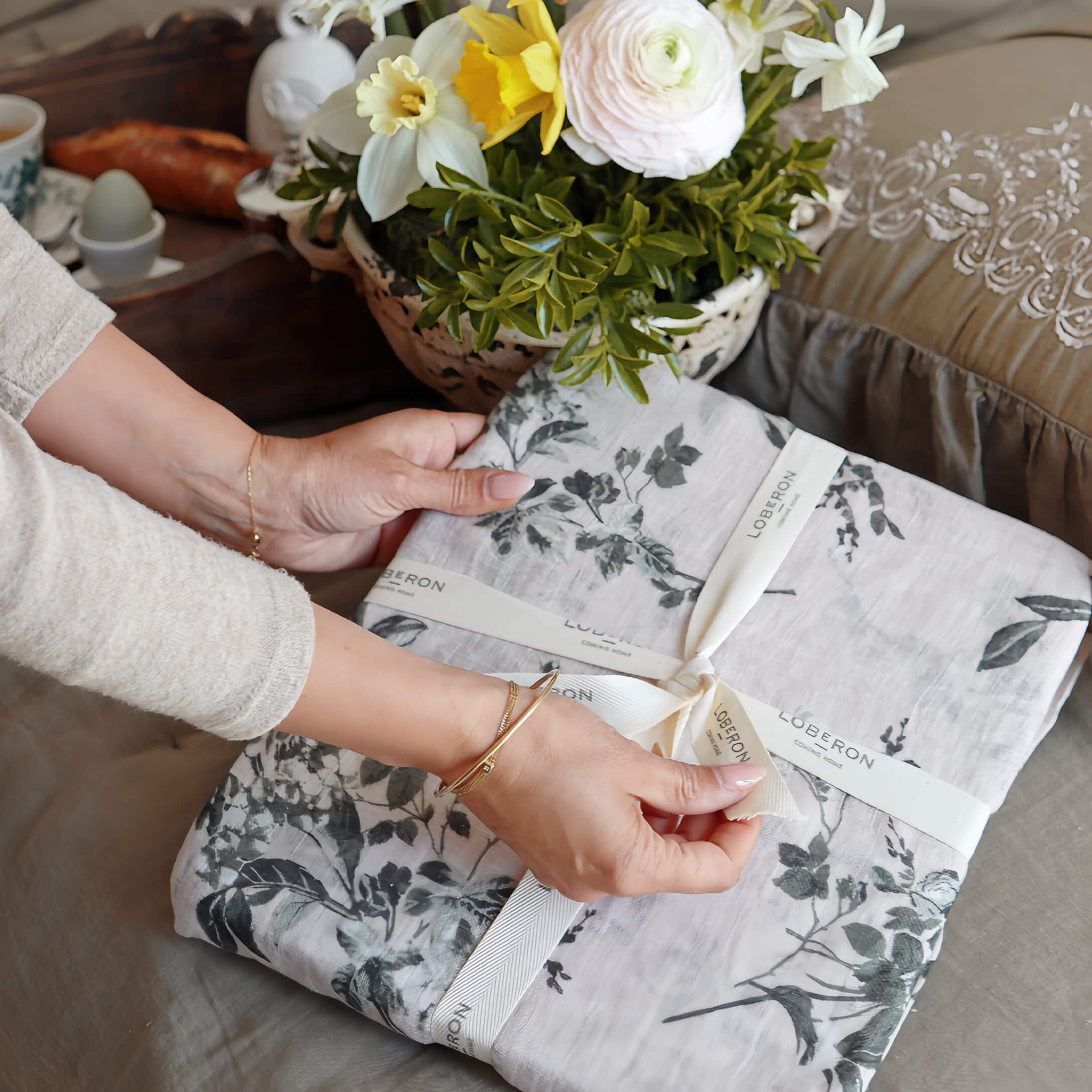 Handen die grijs linnen beddengoed met plantenpatroon uitpakken, bloemenarrangement op tafel