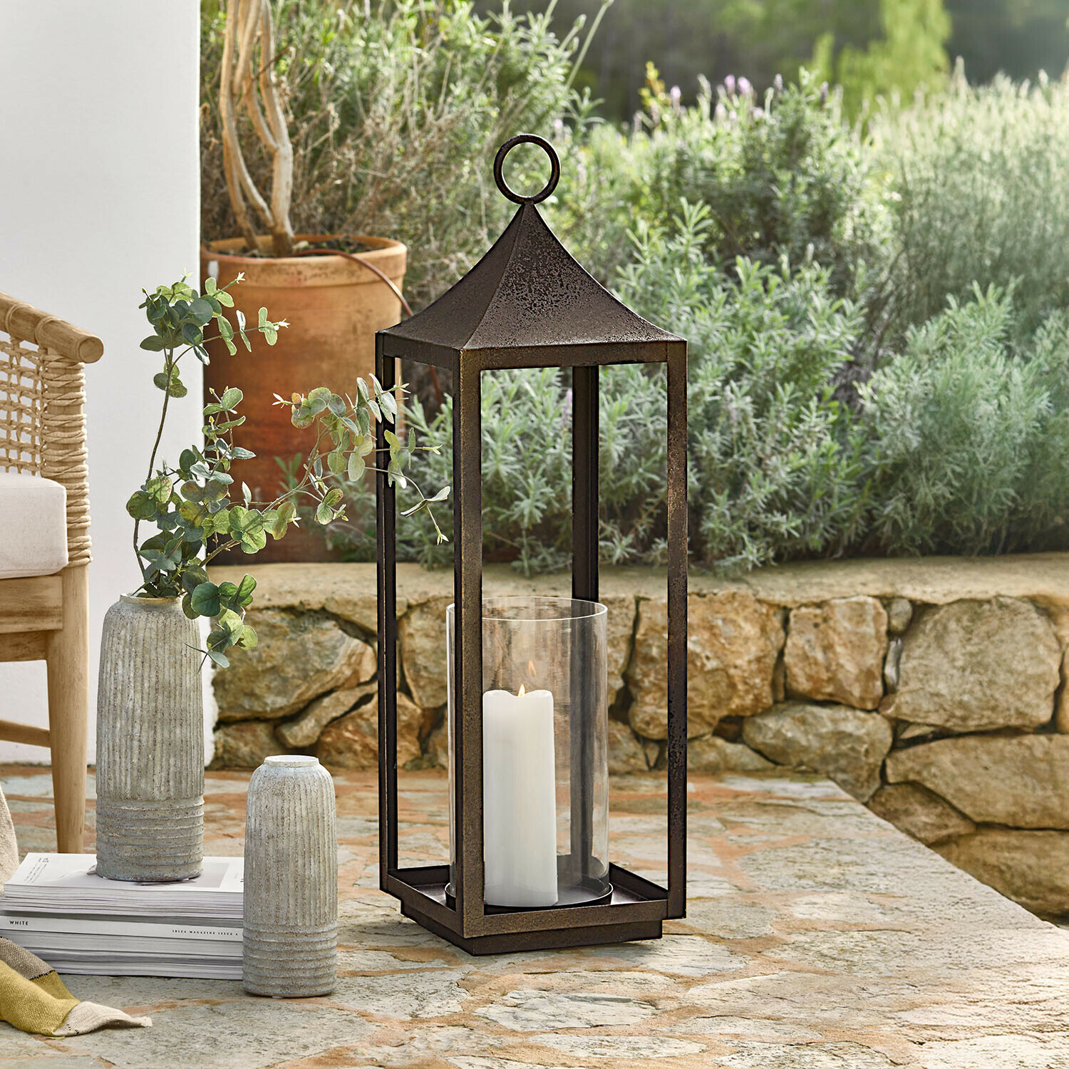 Metalen tuinlantaarn met roestkleur, witte kaars en glazen omhulsel op stenen tafel