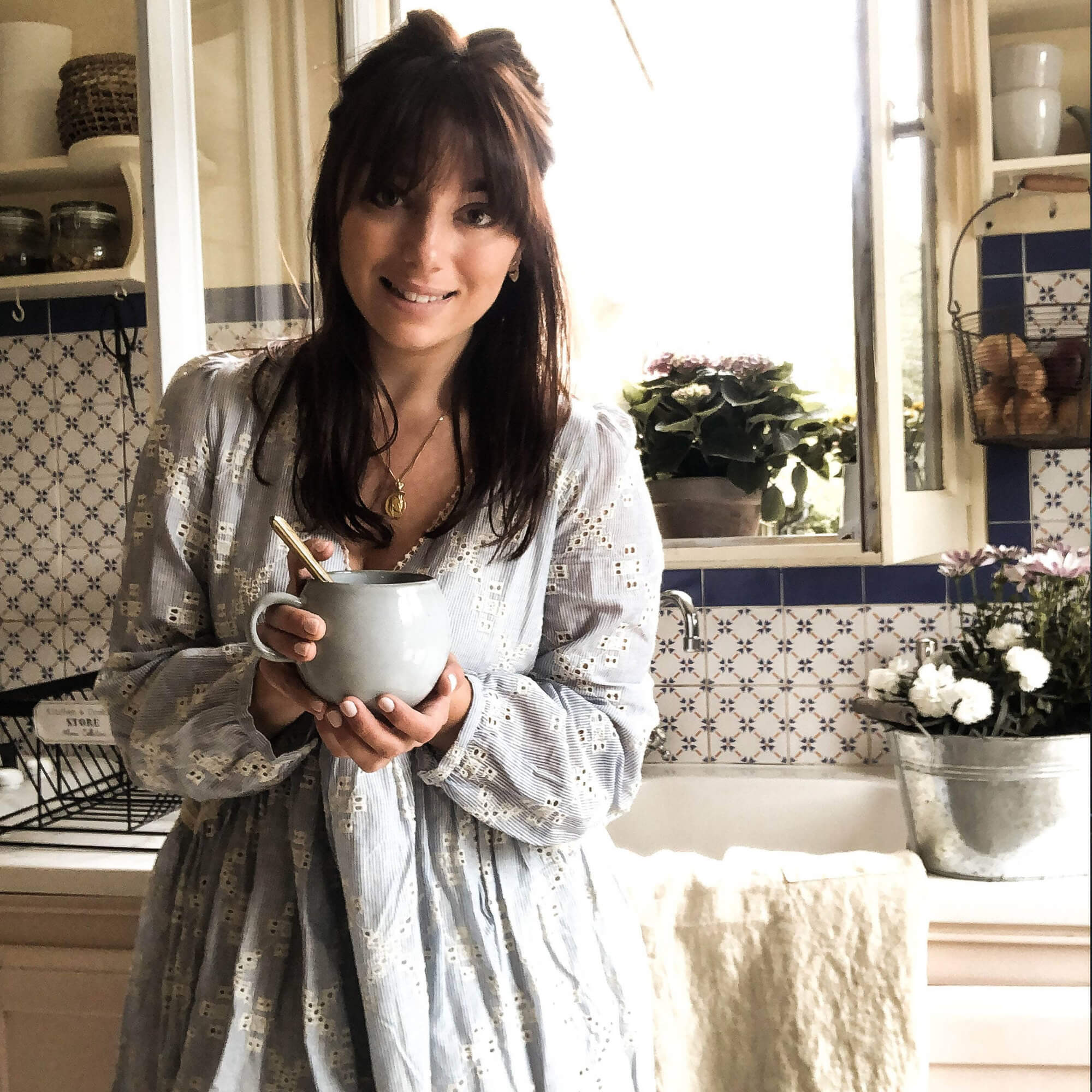 Vrouw in lichte jurk met keramische mok in keuken met blauw-witte tegels en bloemen
