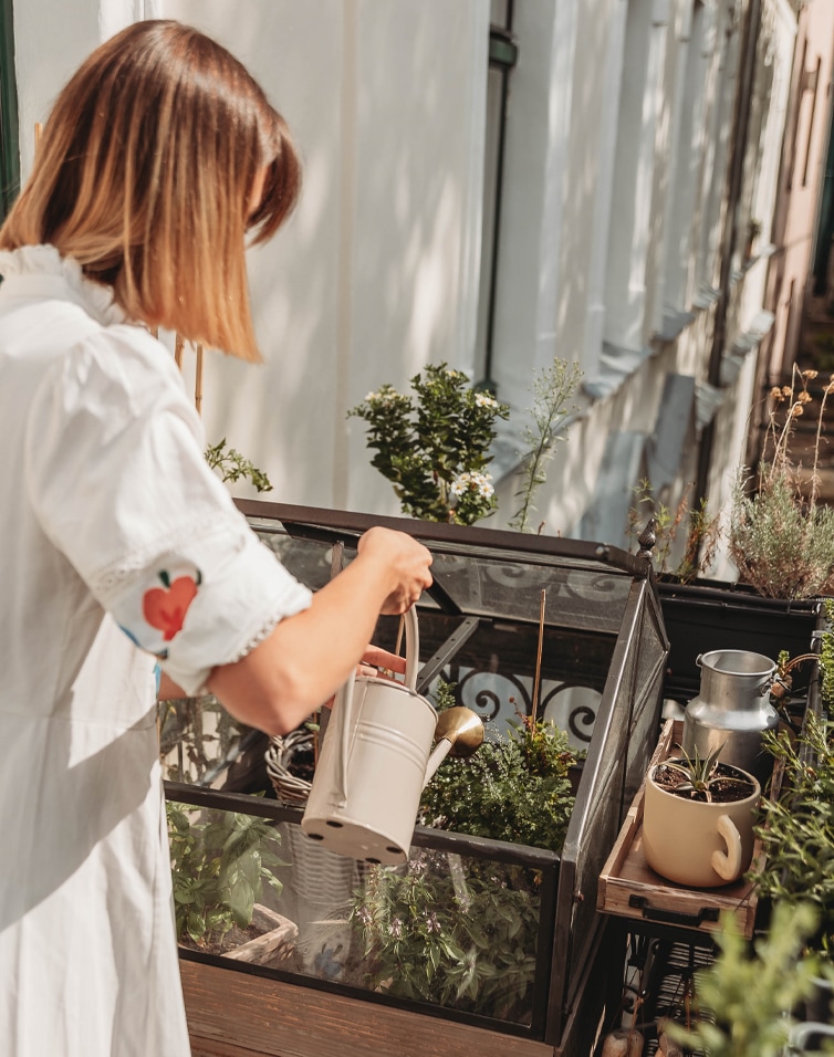 Vrouw die planten water geeft in een kleine glazen kas op een zonnig balkon