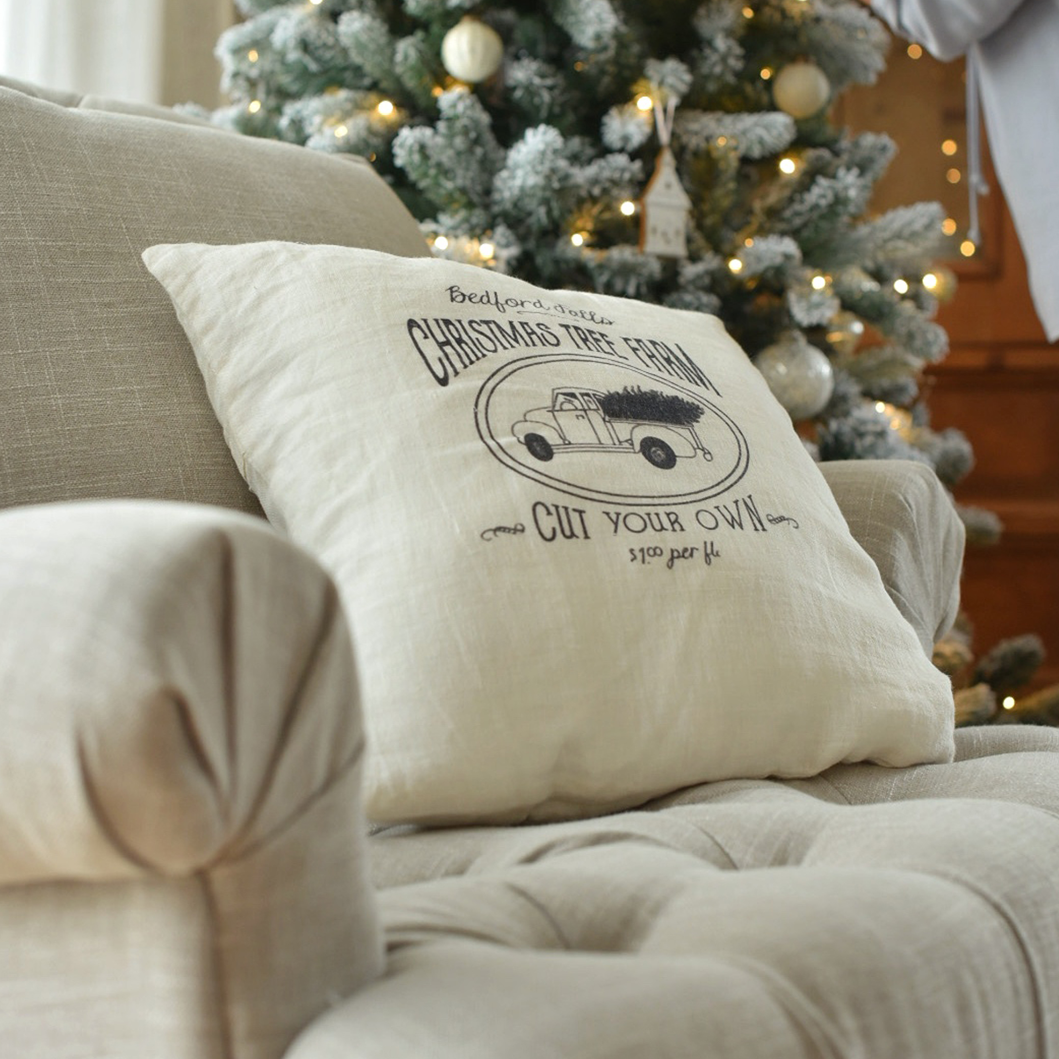 Beige linnen kussen met kersttruckprint op bank bij versierde kerstboom met lichtjes