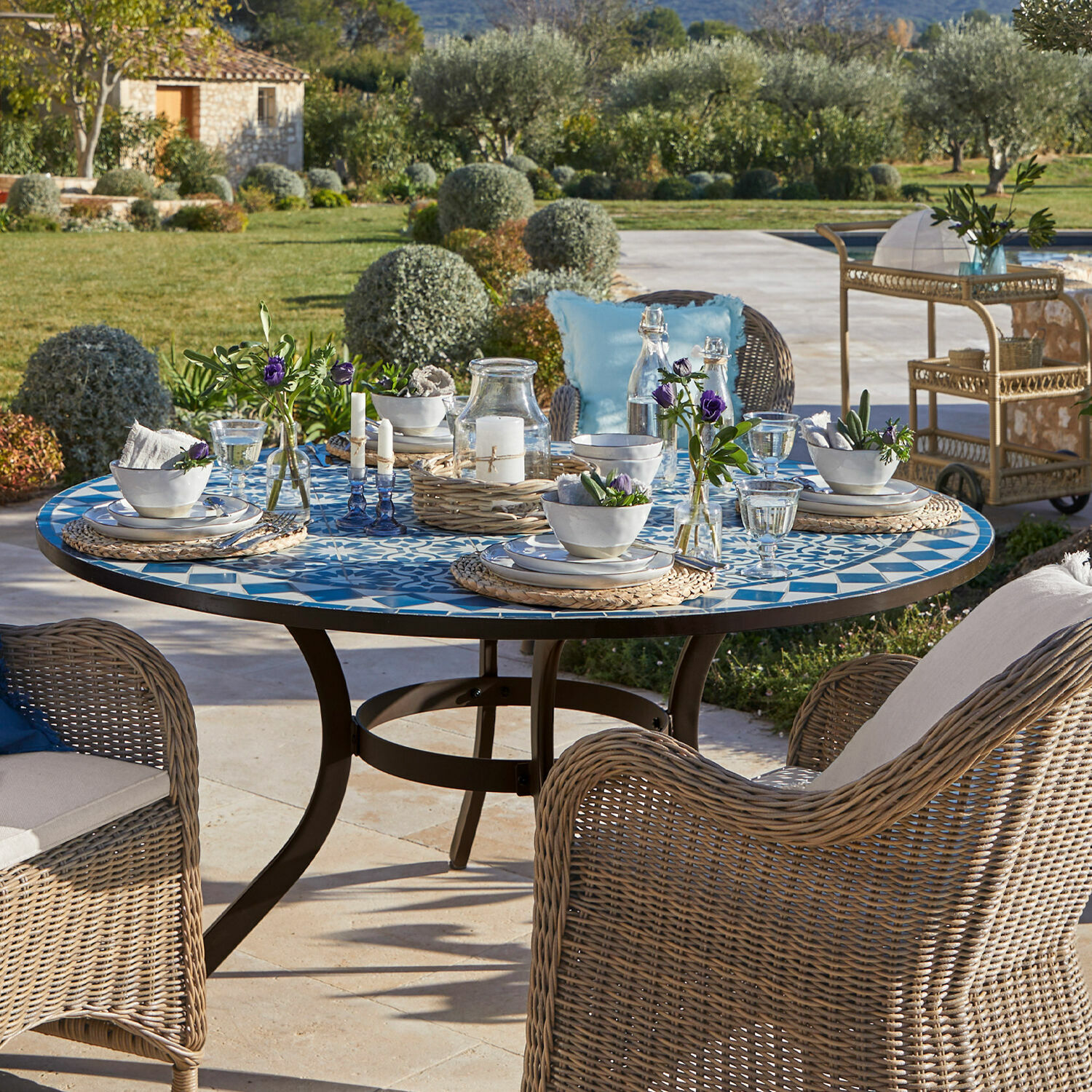 Tafel Jennarton Ronde tuintafel met blauw-wit mozaïekblad en rotan stoelen op het terras