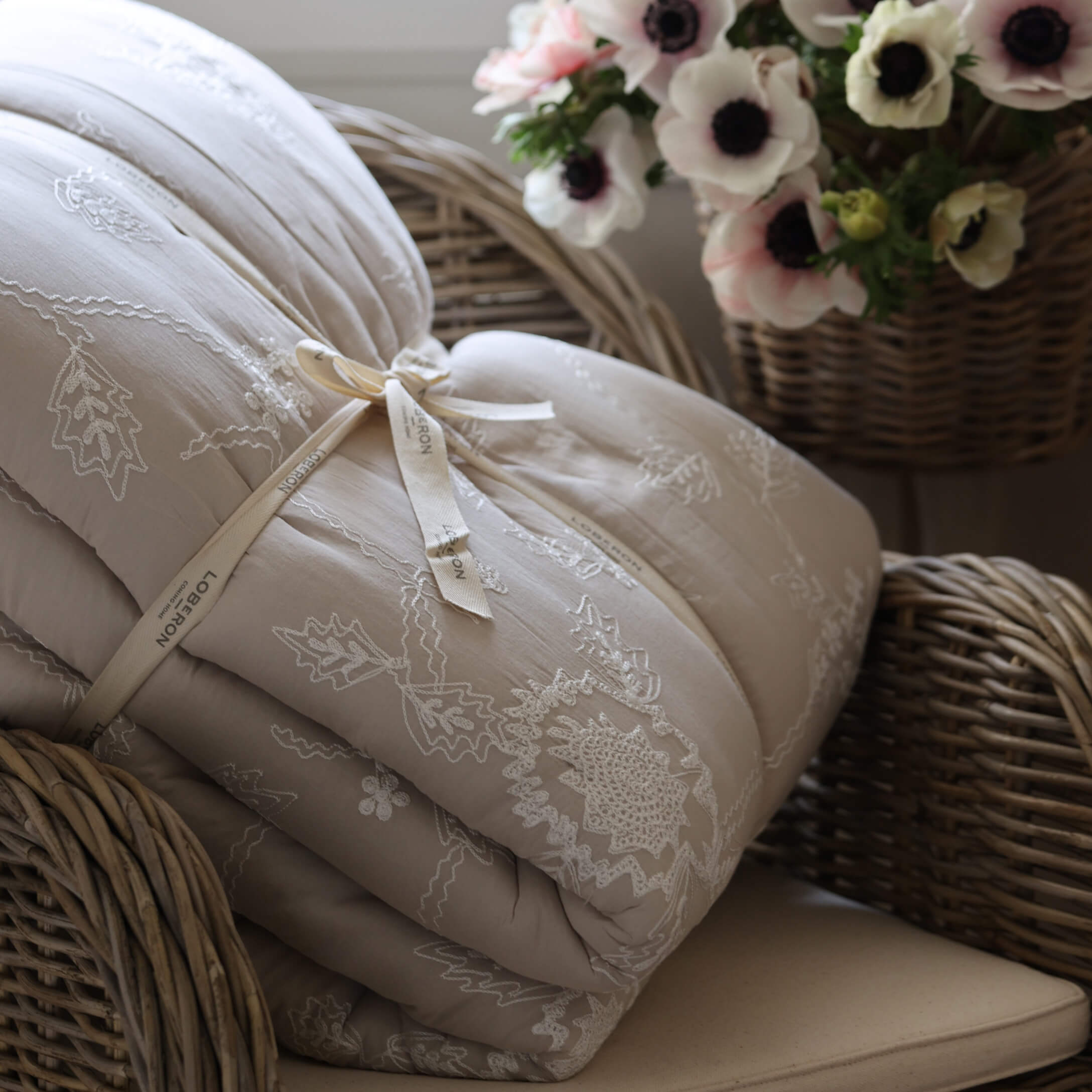 Beige gewatteerde deken met borduurwerk op een rotan stoel naast een bloemenmand, sfeervolle woondecoratie