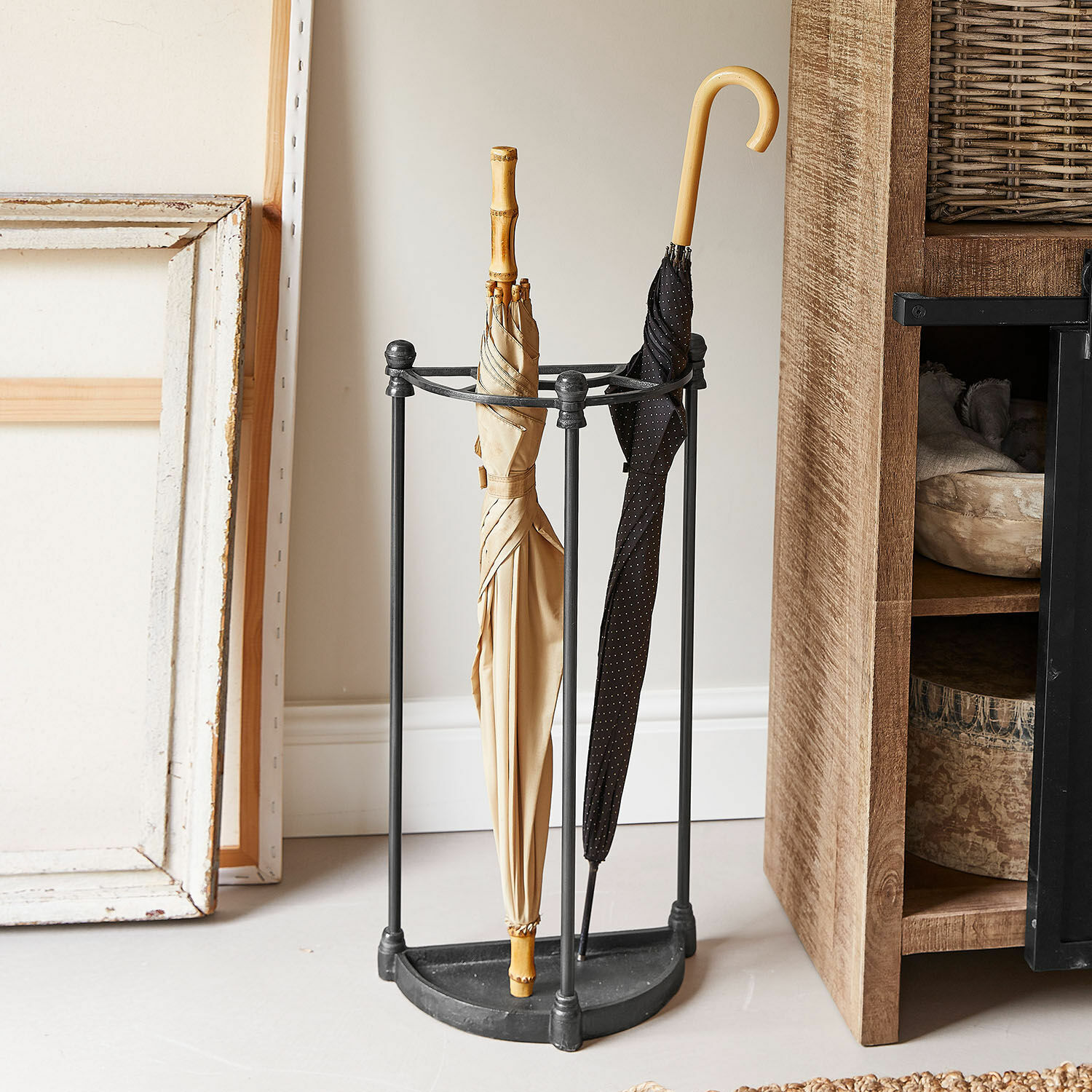 Zwarte metalen paraplubak met twee beige en zwarte paraplu's in een hoek van de kamer