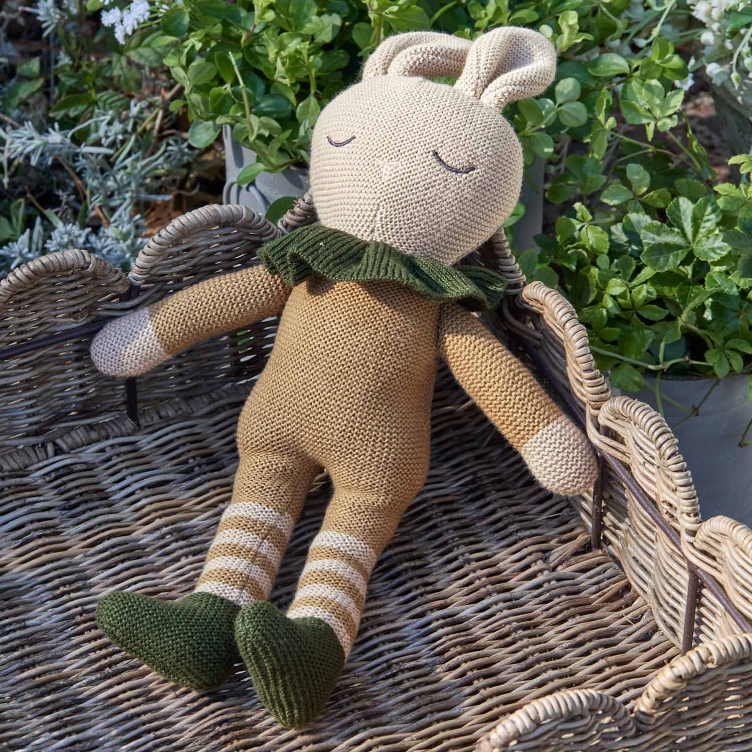 Beige gebreide konijnenknuffel met groene schoenen en kraag op een rieten achtergrond, kinderdecoratie