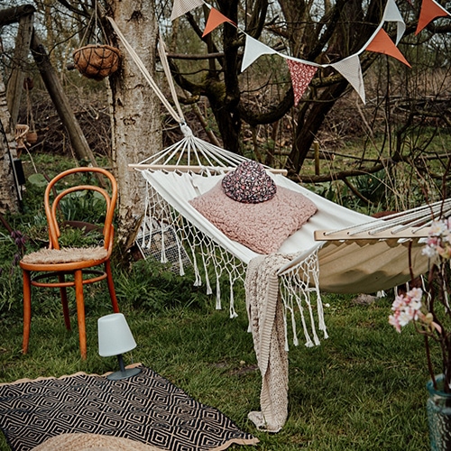 Witte stoffen hangmat met roze kussen en deken op groen gras, houten stoel en decoratieve slinger