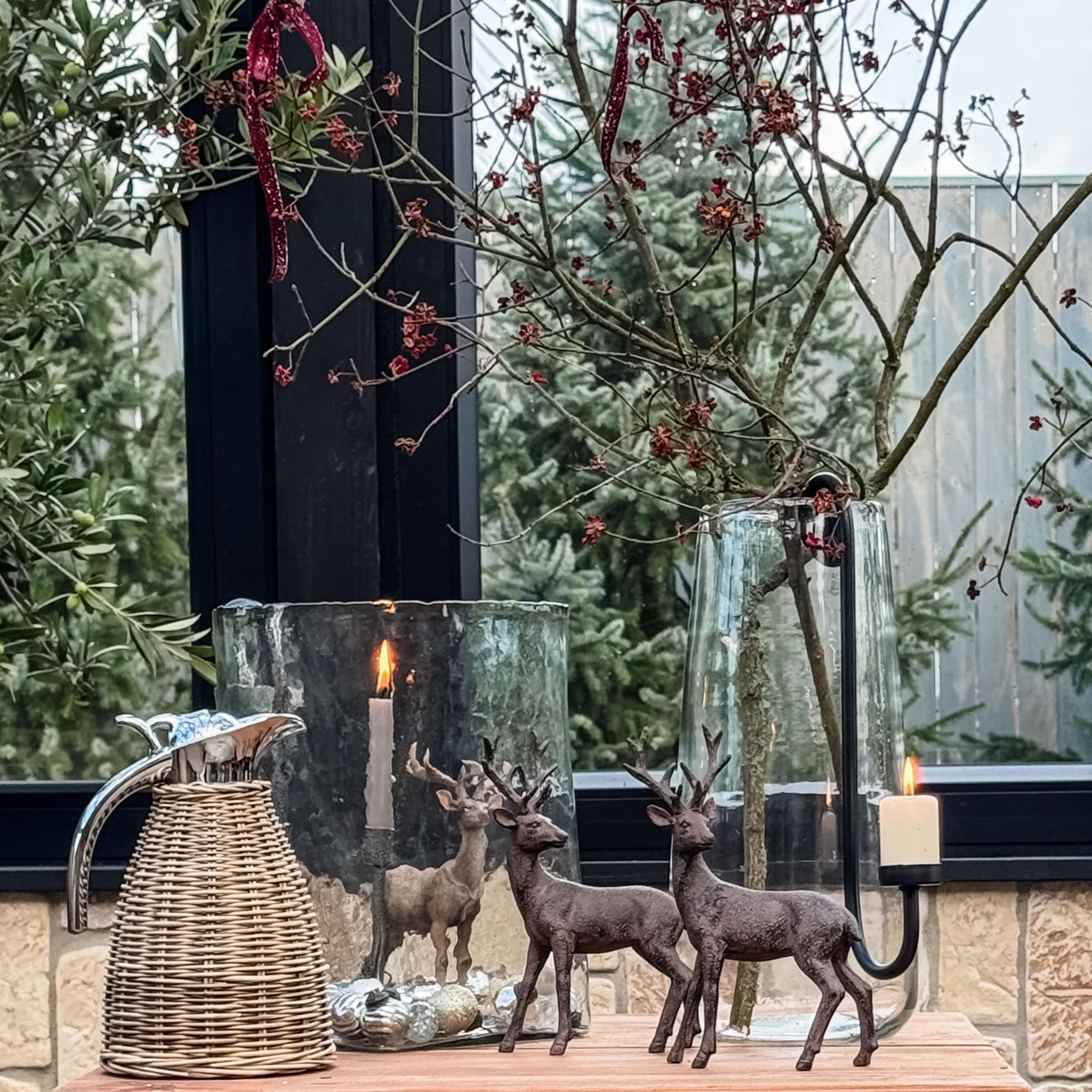 Kerstdecoratie met twee bruine hertenfiguren, brandende kaars en glazen vaas op houten tafel