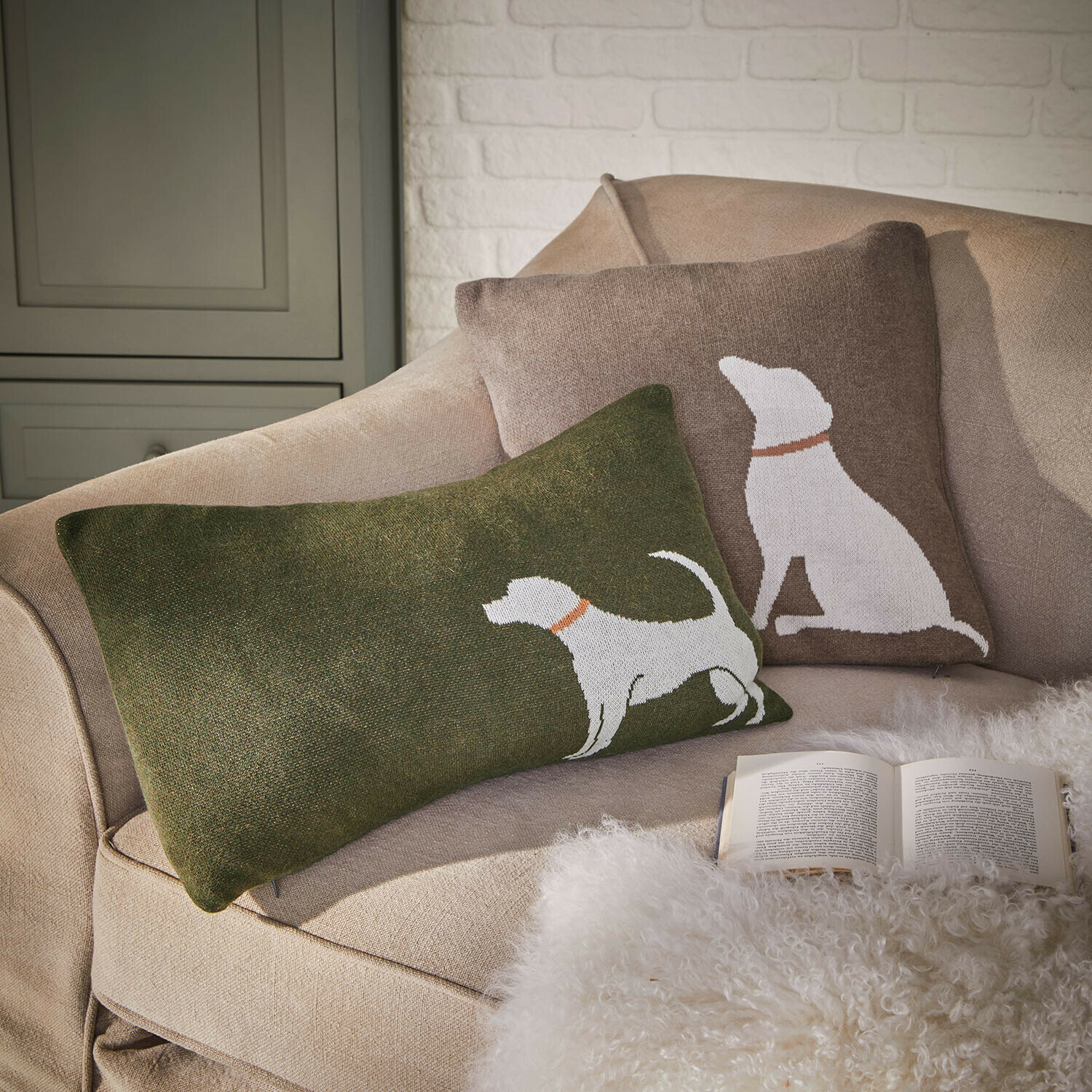 Kussenhoes set van 2 Olyden Set van twee decoratieve kussens met wit hondenmotief op groene en bruine achtergrond op een beige bank
