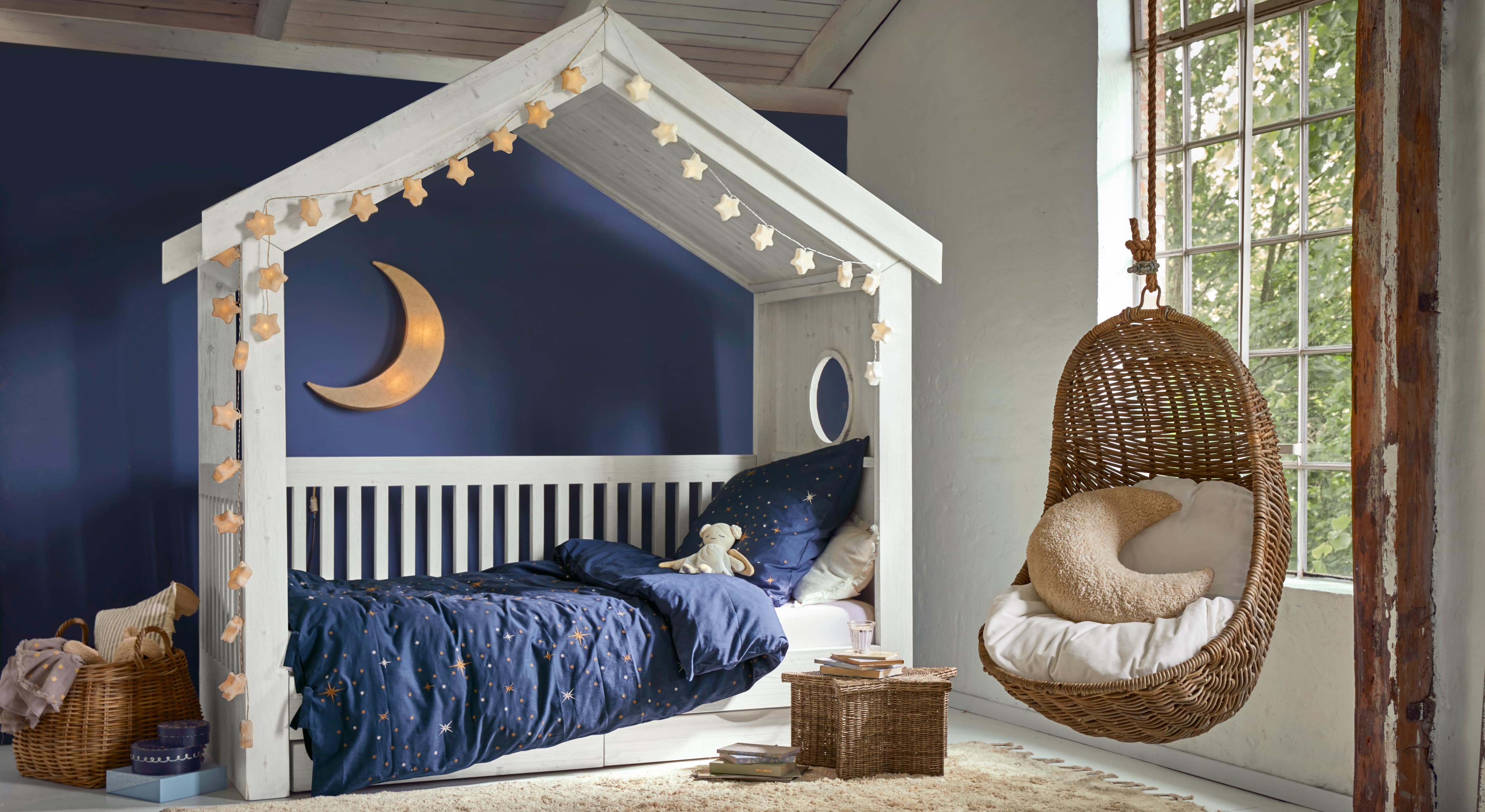 Wit houten huisvormig bed met donkerblauw sterrenbeddengoed en hangende rotan stoel in kinderkamer
