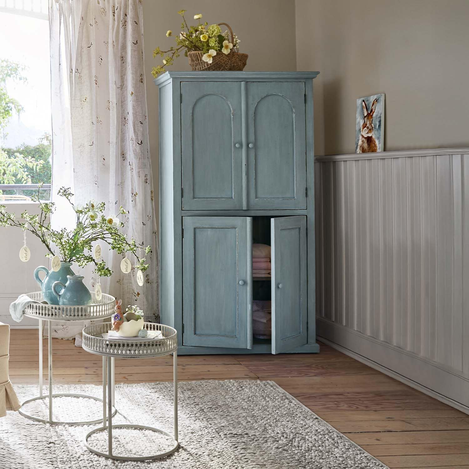 Blauwe houten kast met twee deuren in lichte kamer met bloemen en vloerkleed