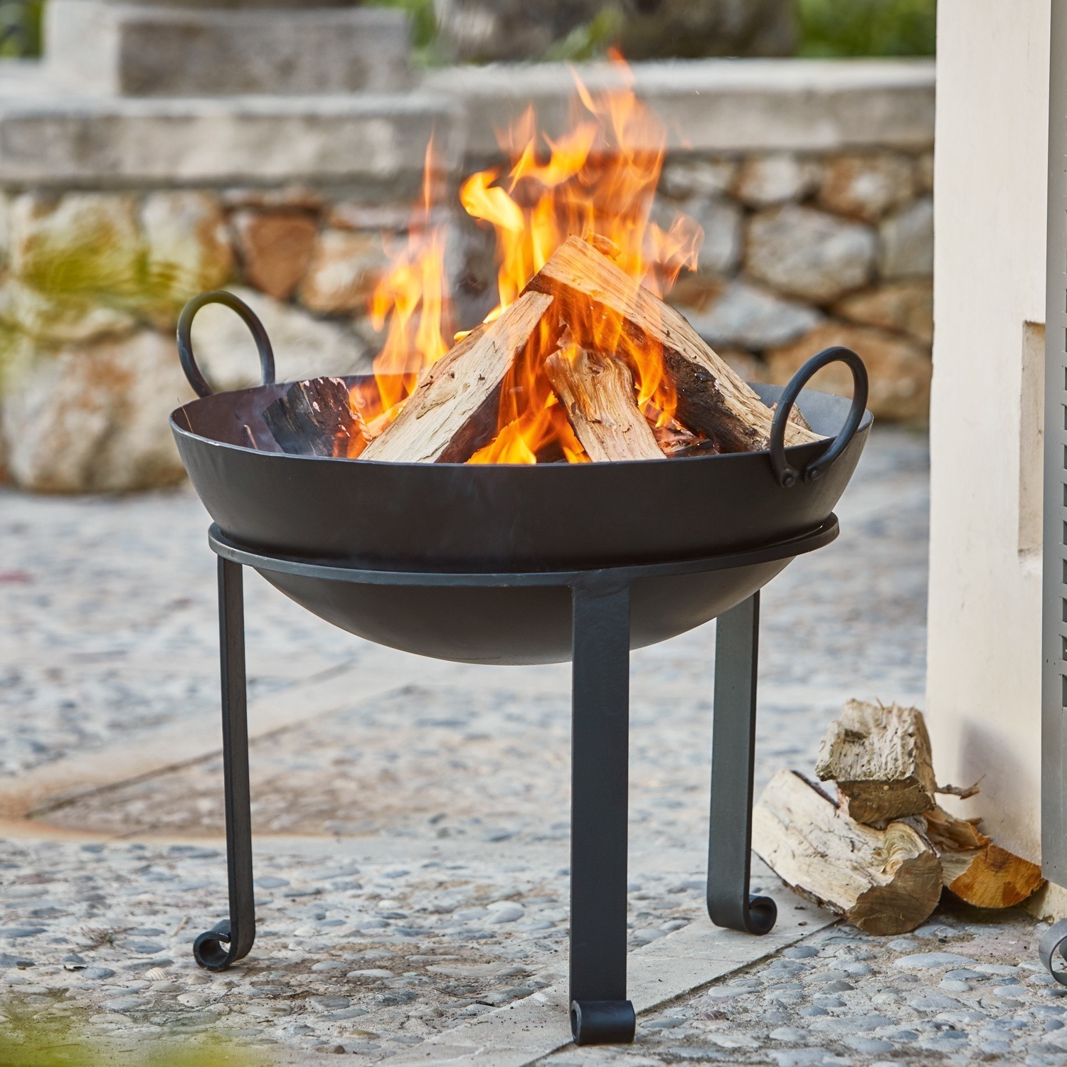 Vuurschaal Harrogate Zwarte metalen vuurkorf met brandend hout op stenen terras buiten