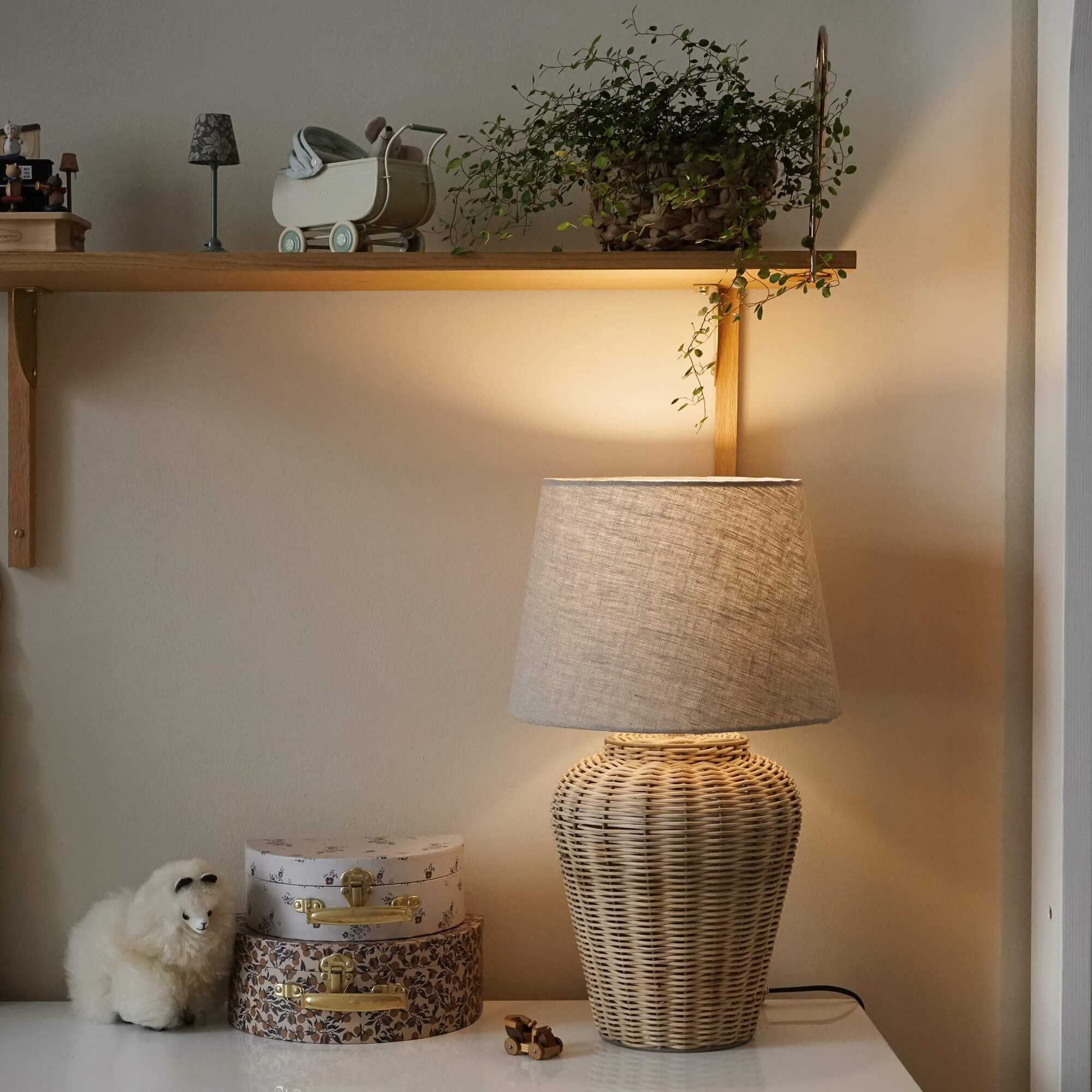 Tafellamp van gevlochten riet met beige kap op wit blad met decoraties en plant op plank
