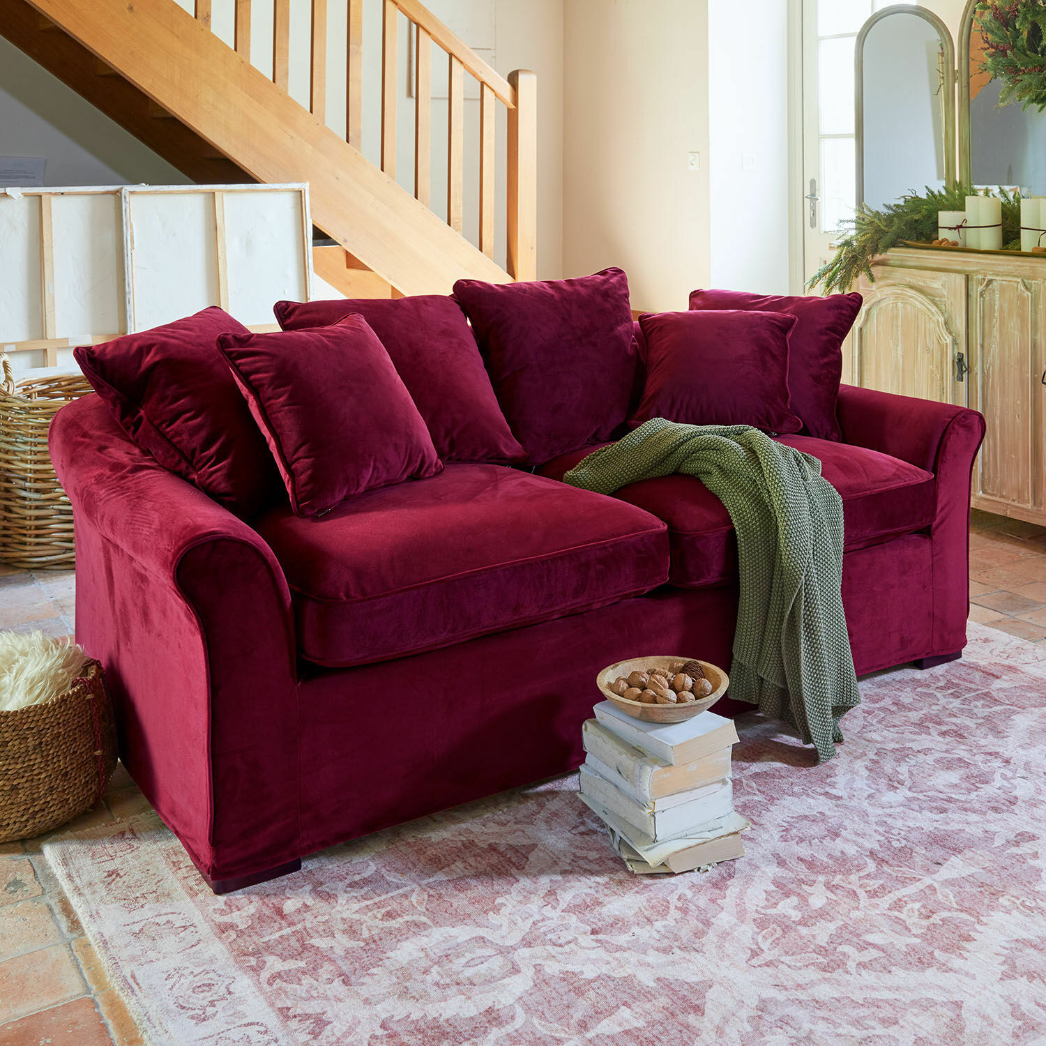 Bordeauxrode fluwelen bank met kussens en groene deken op rustieke vloerkleed in woonkamer
