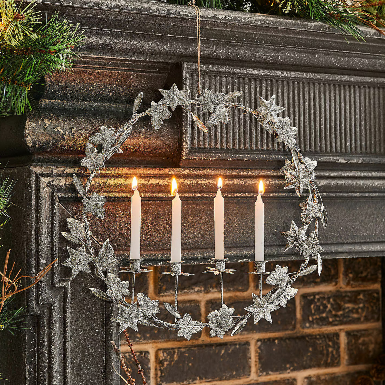 Metalen krans met bladeren en vier brandende witte kaarsen aan een schoorsteen, kerstdecoratie