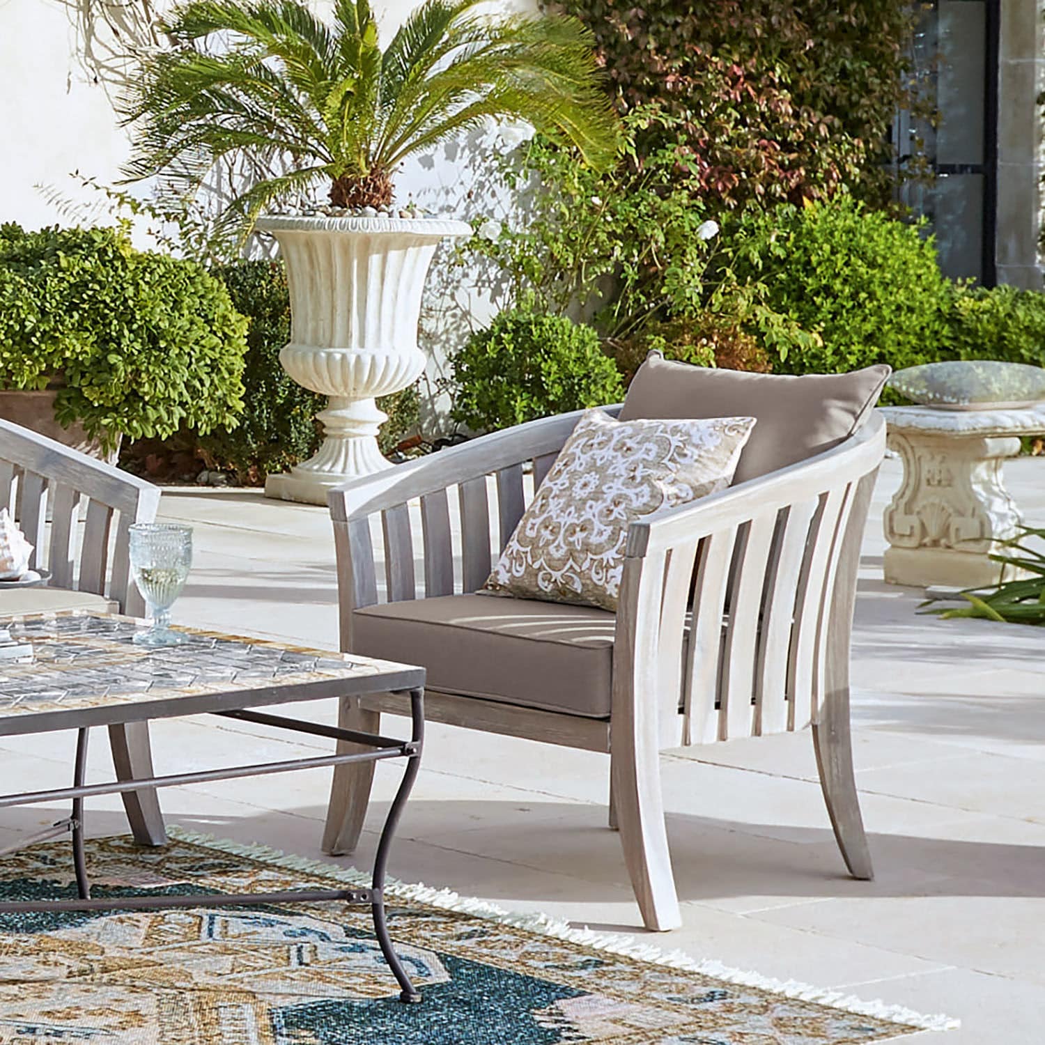 Tuinstoel van licht hout met beige kussens op terras met groene planten op de achtergrond