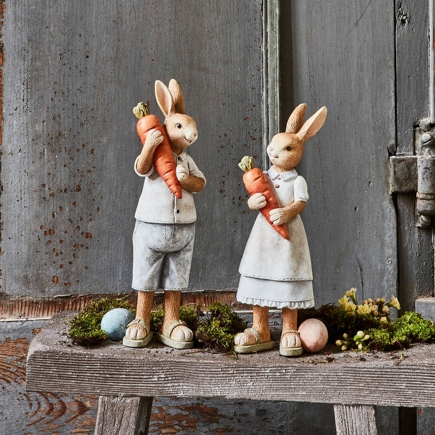 Haas set van 2 Gimont Beeldjes van konijnen van materiaal in beige kleding met oranje wortels op een rustieke houten bank