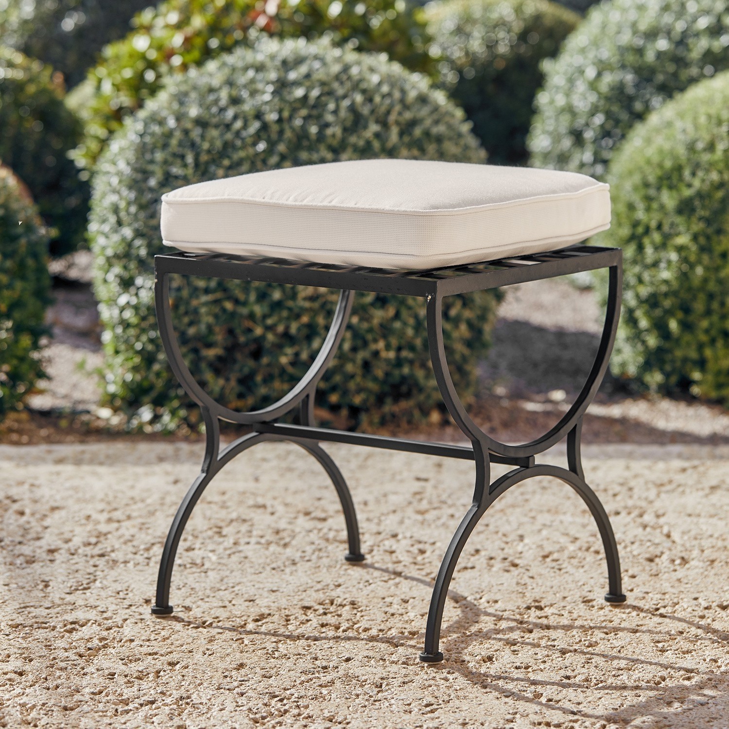 Hocker Terell Metalen tuinkruk met zwart frame en crème kussen op zandige ondergrond buiten