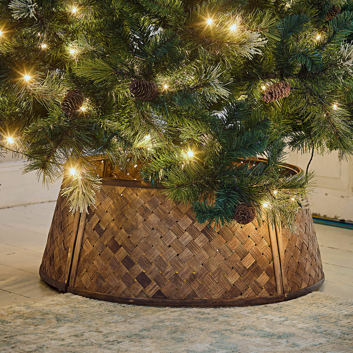 Omranding voor de kerstboom Eden Bruine gevlochten houten kerstboomstandaard onder groene boom met lichtjes en dennenappels