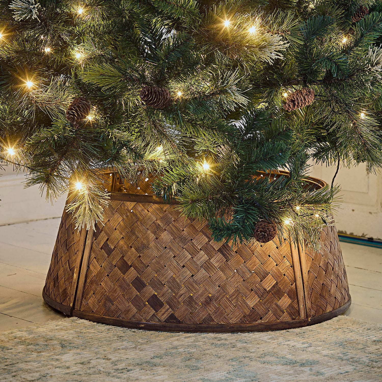 Omranding voor de kerstboom Eden Bruine gevlochten houten kerstboomstandaard onder groene boom met lichtjes en dennenappels