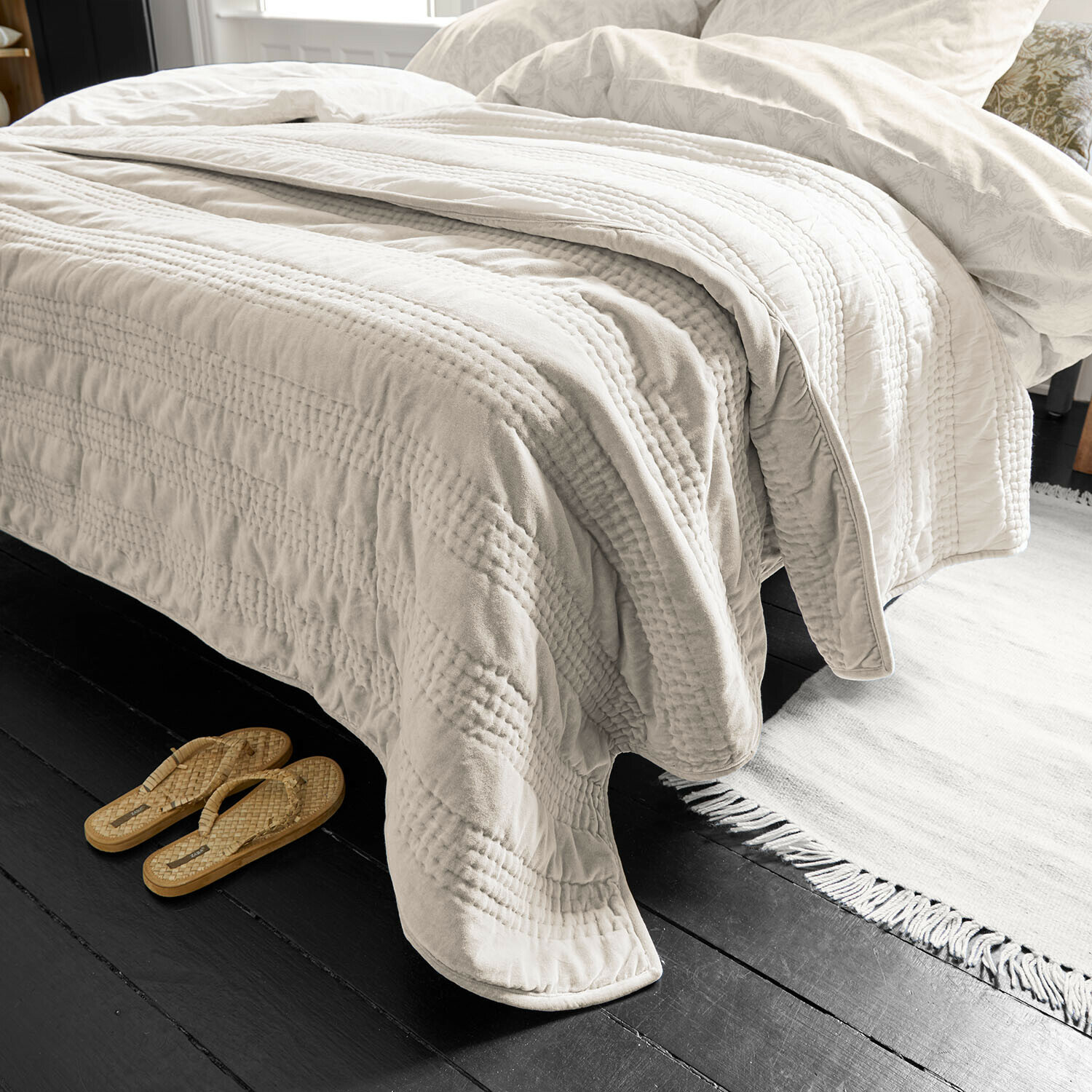 Beige doorgestikte deken van zacht materiaal op bed met lichte beddengoed en slippers ernaast