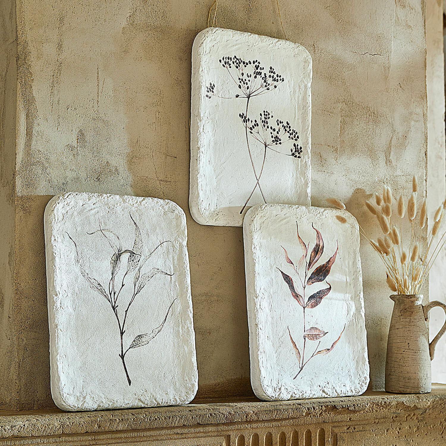 Afbeelding set van 3 Hillsdale Drie witte wanddecoraties met plantenmotieven aan een beige muur, natuurlijke rustieke stijl.