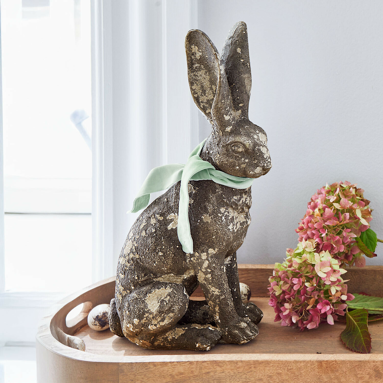 Decoratieve haas van gepatineerd steen met groene strik op houten dienblad met bloemen