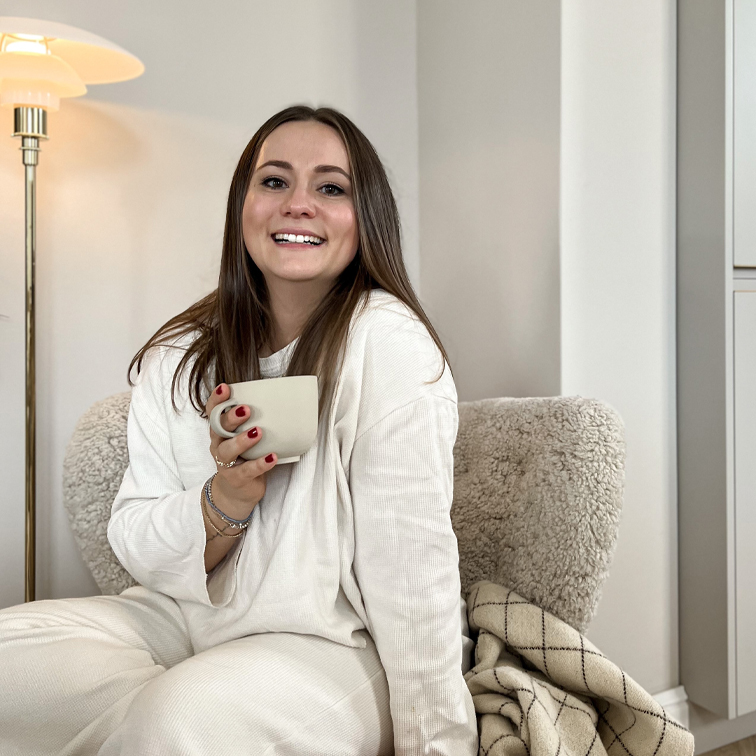 Lachende vrouw in zachte witte kleding zittend op stoel met mok in gezellige kamer