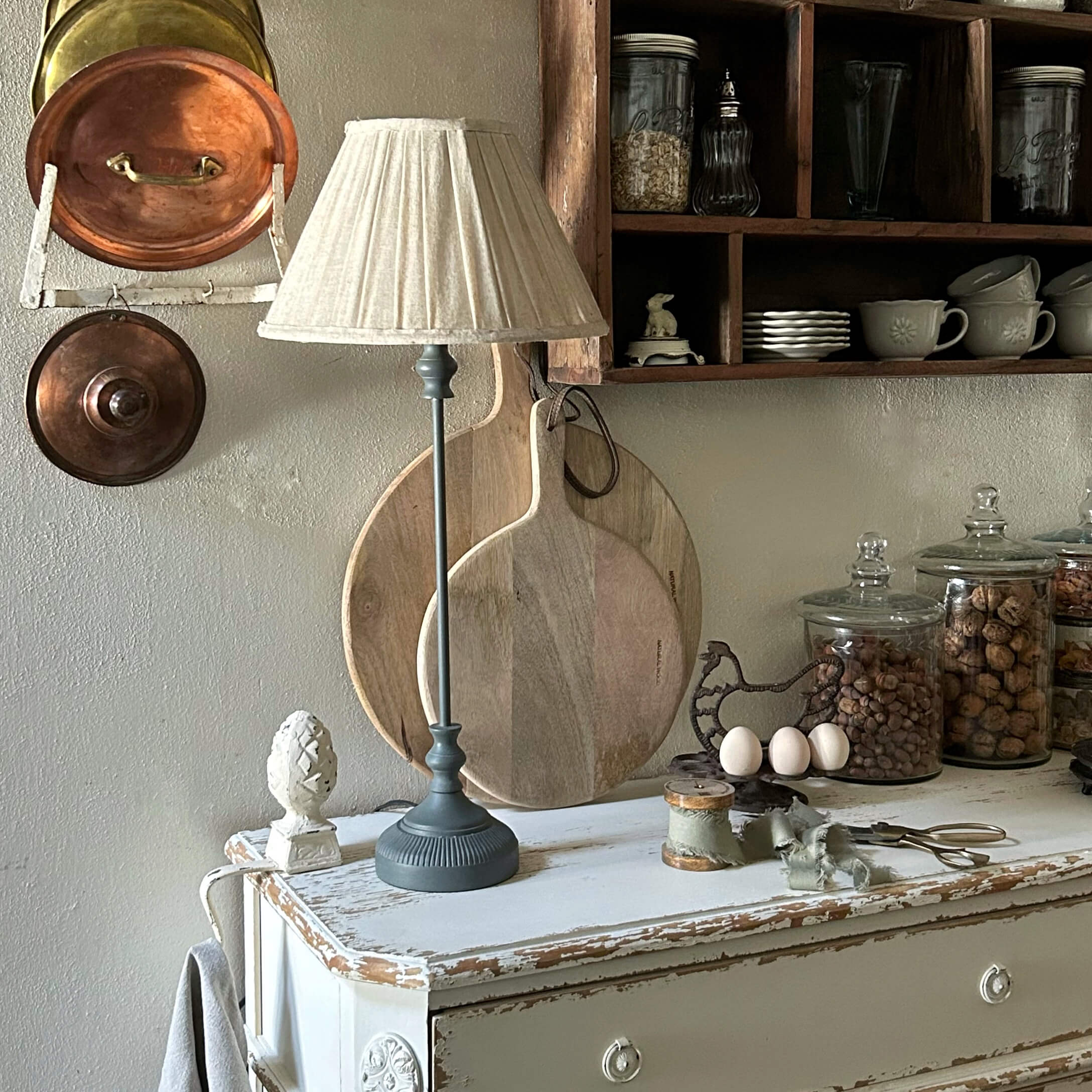 Tafellamp met grijze voet en crèmekleurige kap op een verweerde witte kast in een rustieke keuken