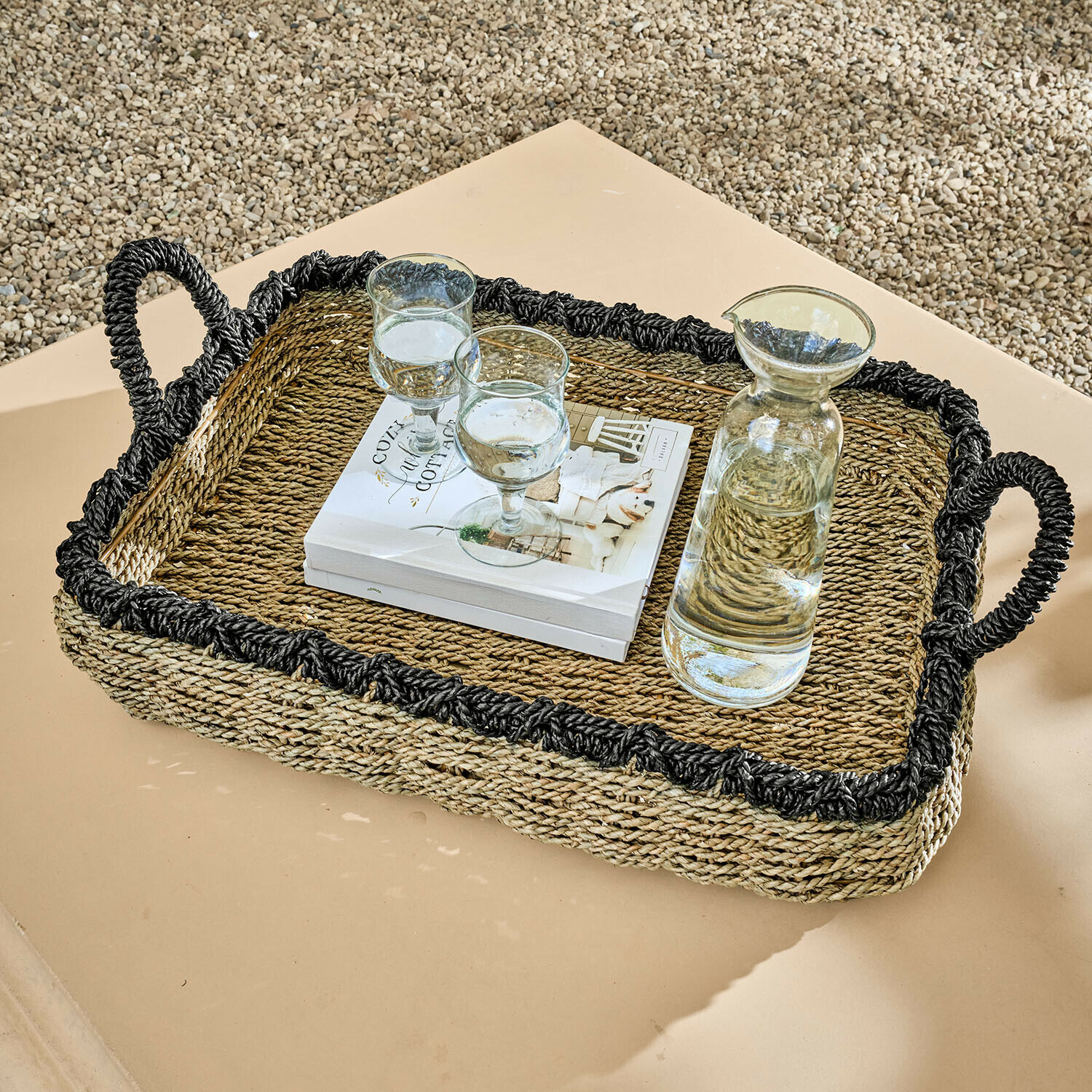 Rechthoekige dienblad van gevlochten zeegras met zwarte rand en handvatten, met karaf en glazen op tafel