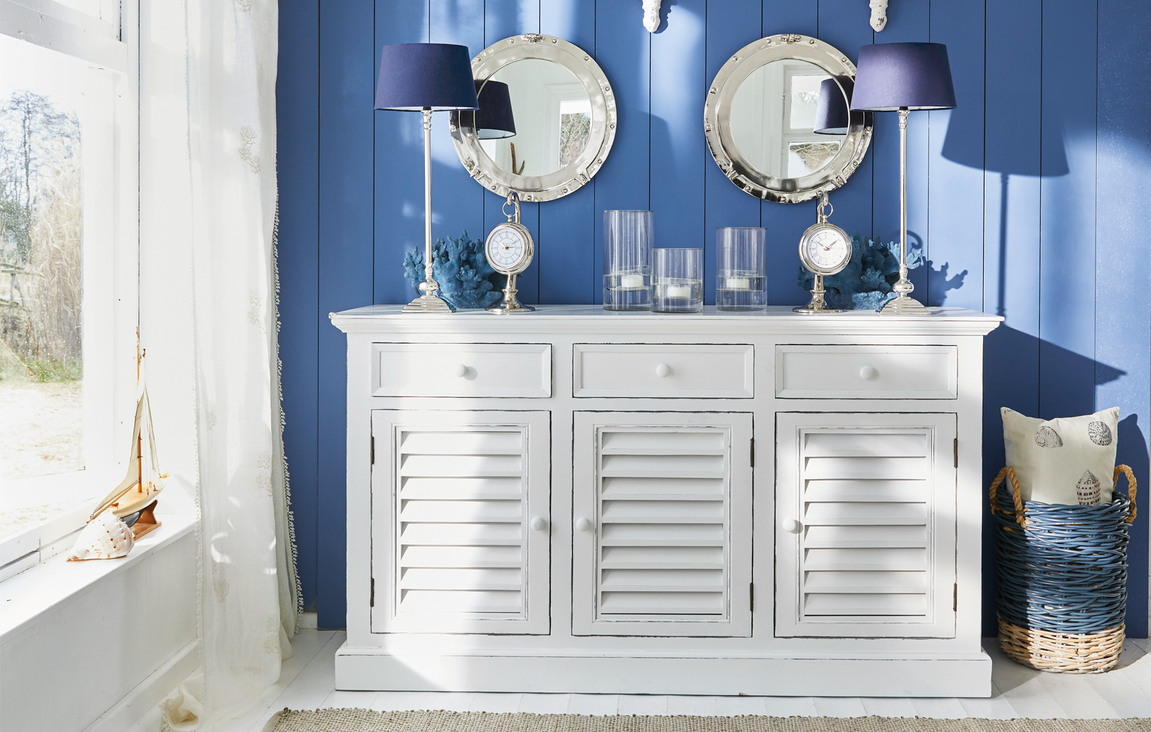 Witte houten dressoir met lamellen deuren tegen blauwe muur met maritieme decoratie