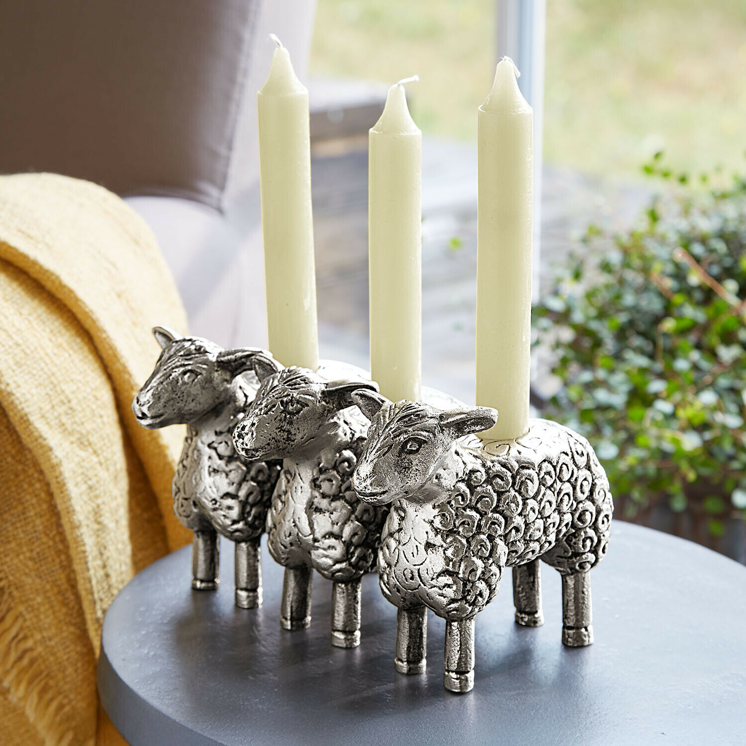Drie zilverkleurige kaarsenhouders in schapenvorm met witte kaarsen op een grijze tafel